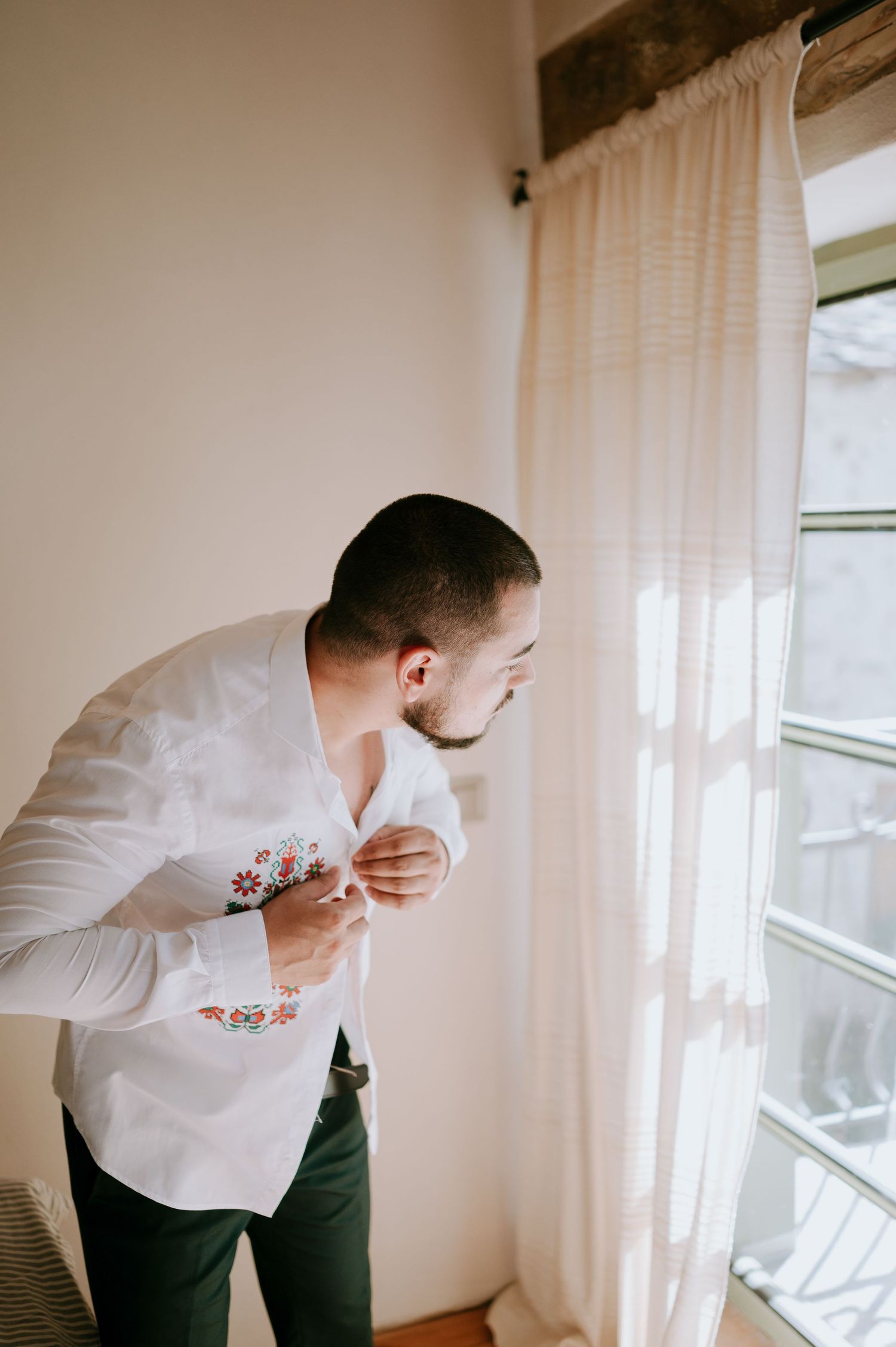 Person in white embroidered shirt buttoning up near a window with sheer curtains.