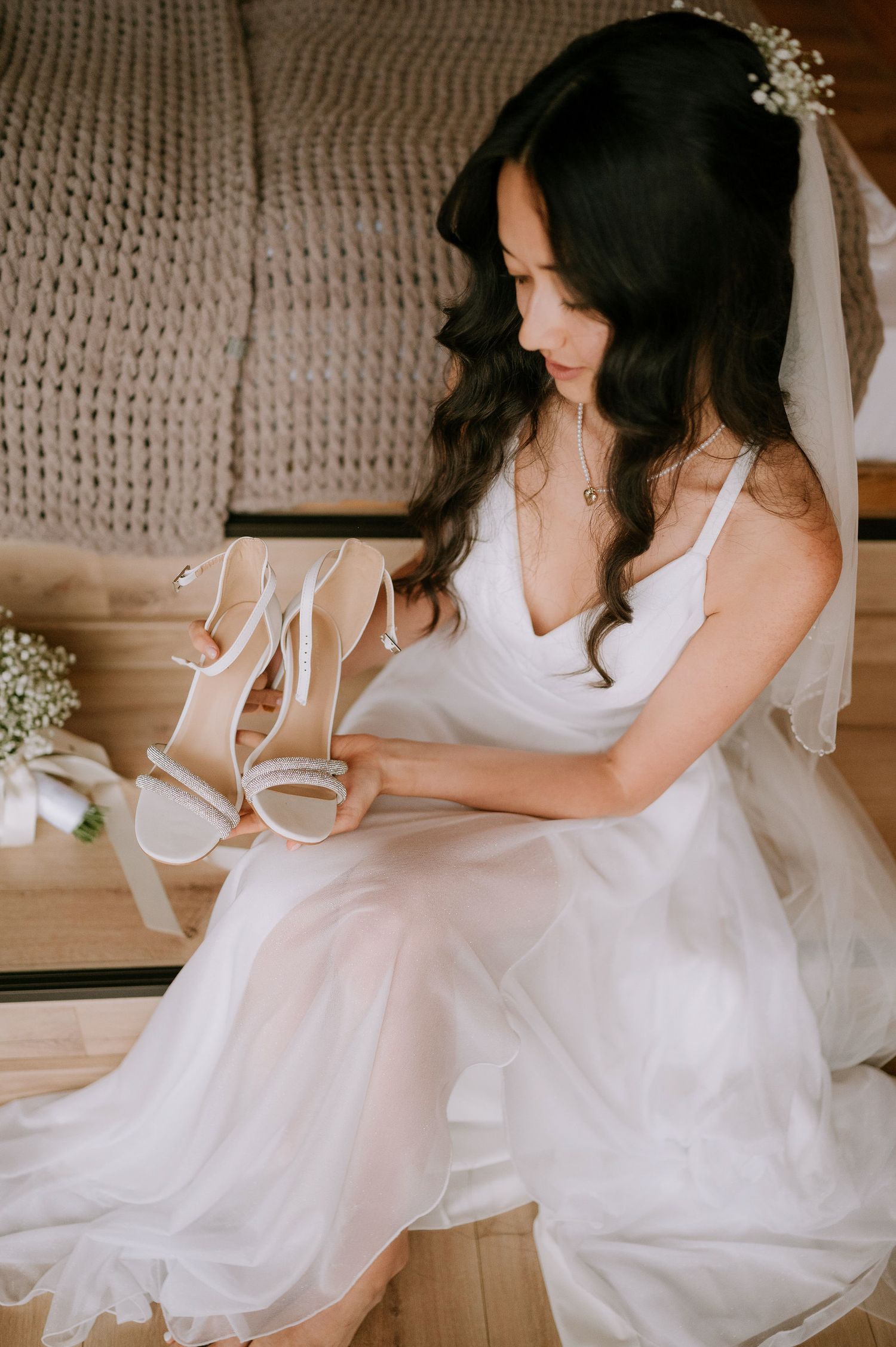 Bride in flowing white dress admires wedding shoes while sitting in rustic setting.