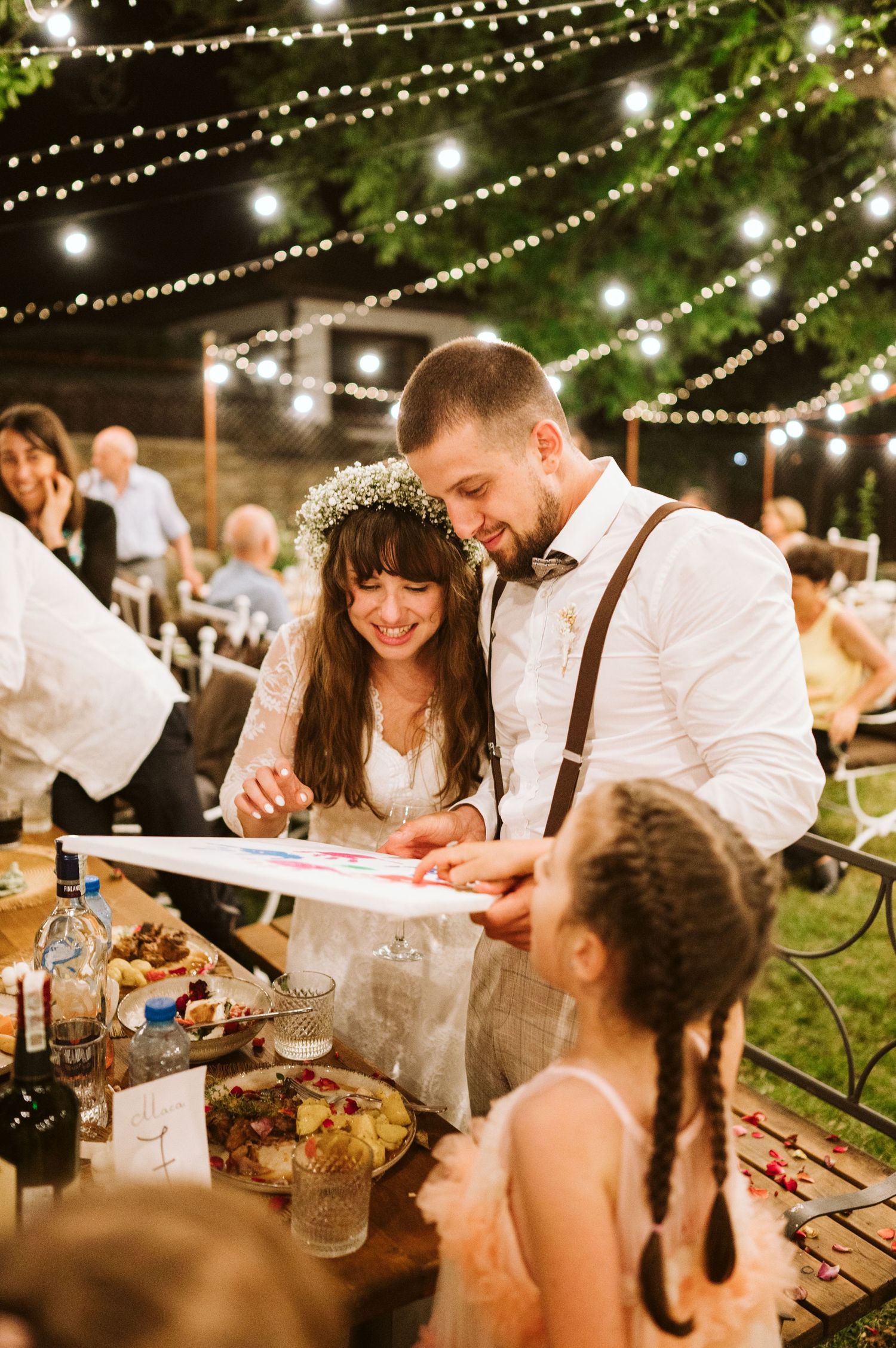 A wedding celebration at night under string lights with guests enjoying food and drinks.
