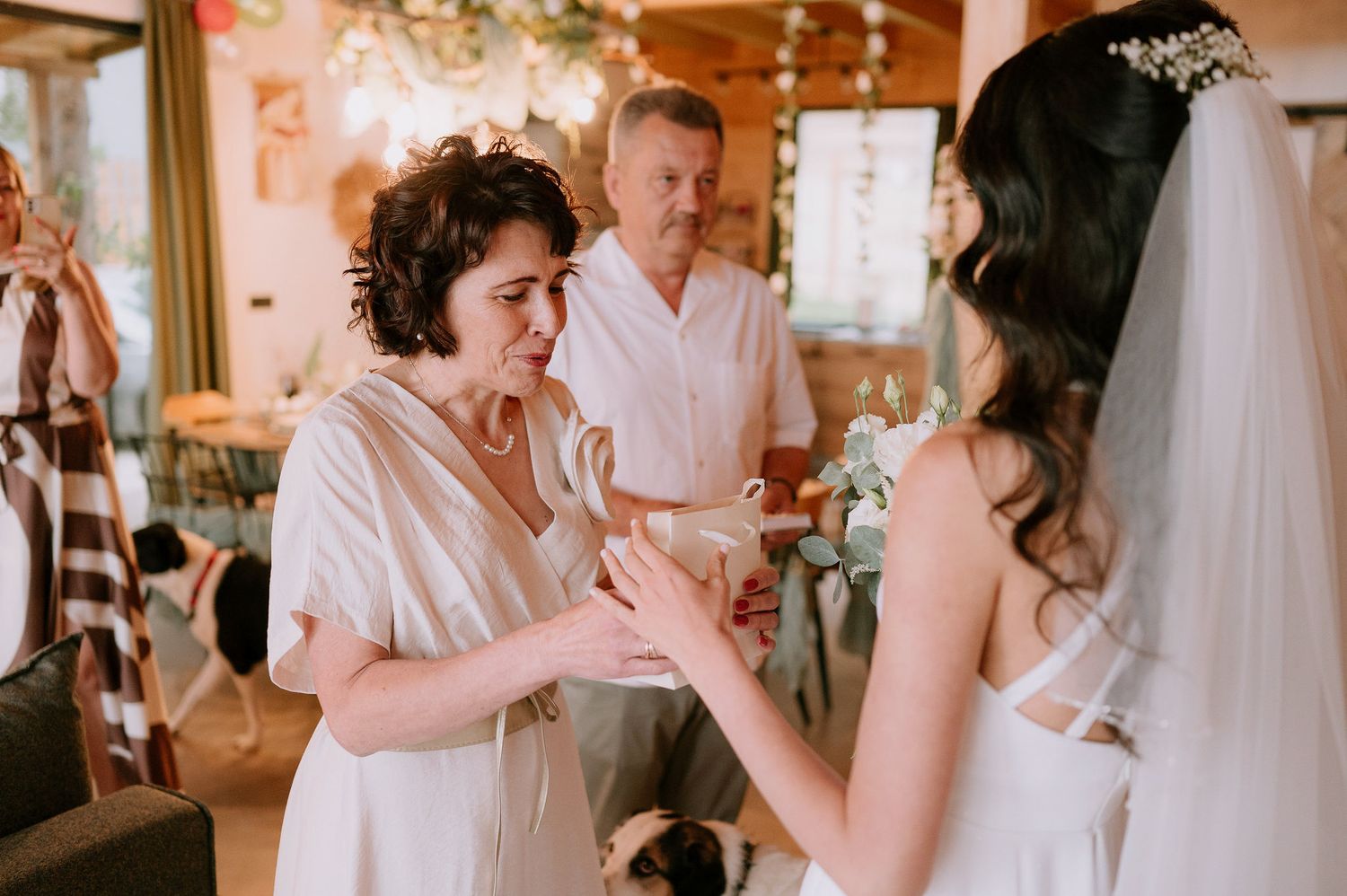 Wedding guests interact during a reception in a warmly lit wooden interior with hanging decorations.