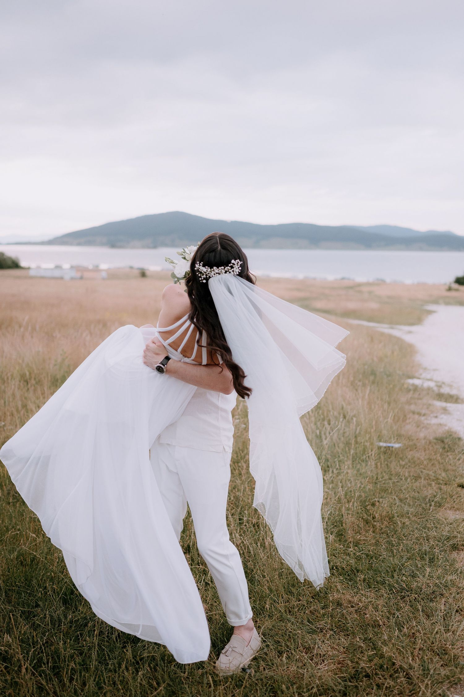 A couple in wedding attire dances playfully in a grassy field with mountains and water in the background.