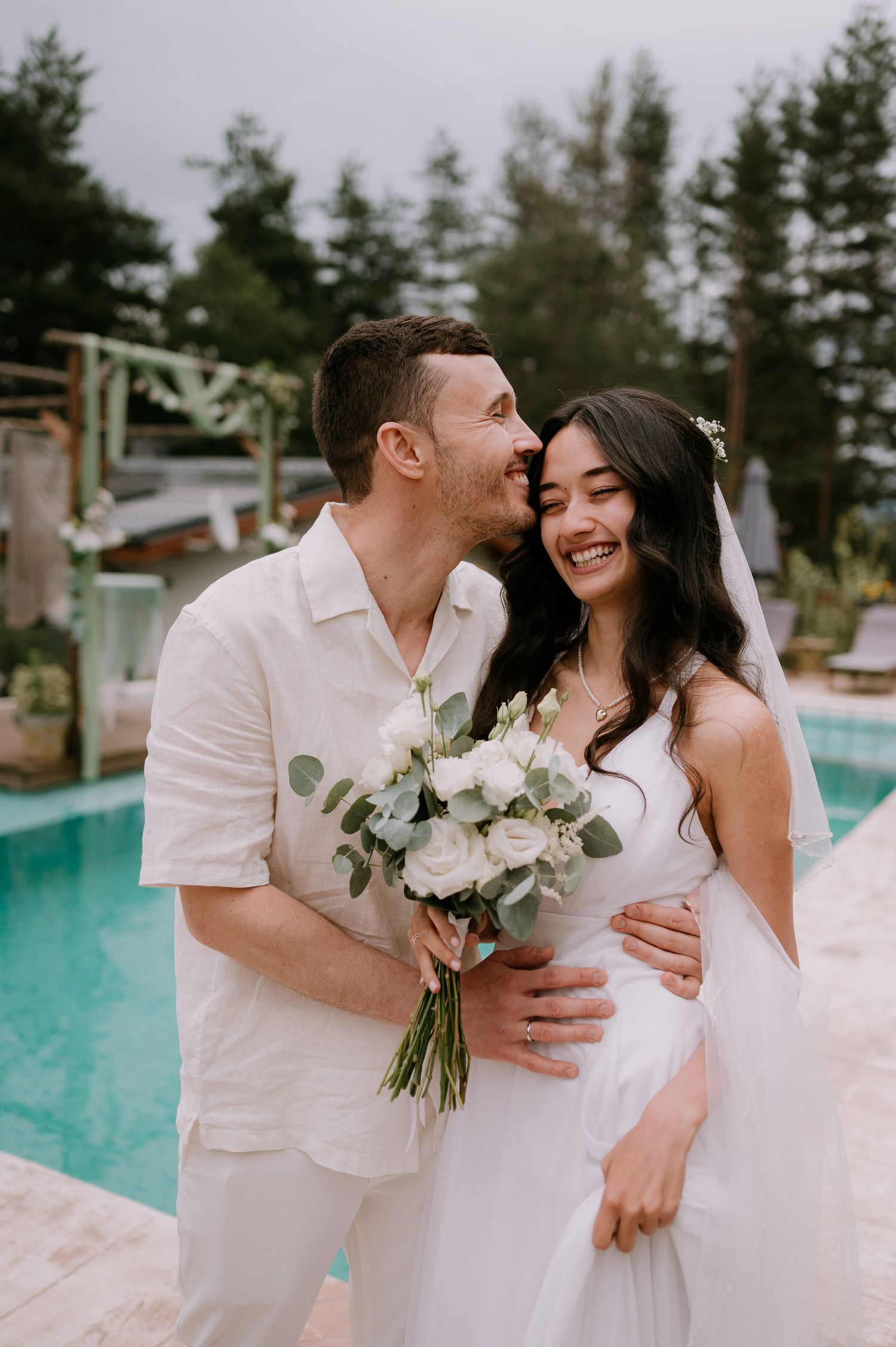 Happy couple laughs together during their white-themed poolside wedding photoshoot.