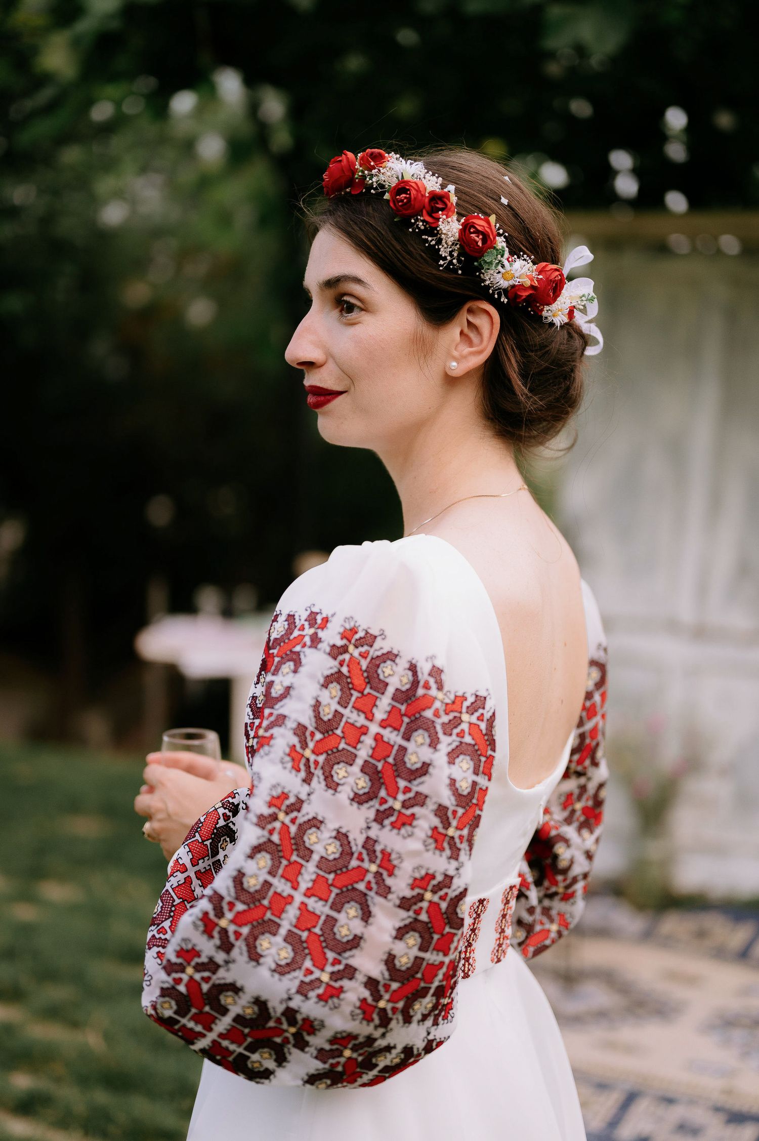 Bride wearing white dress with red embroidered sleeves and floral crown in outdoor setting.