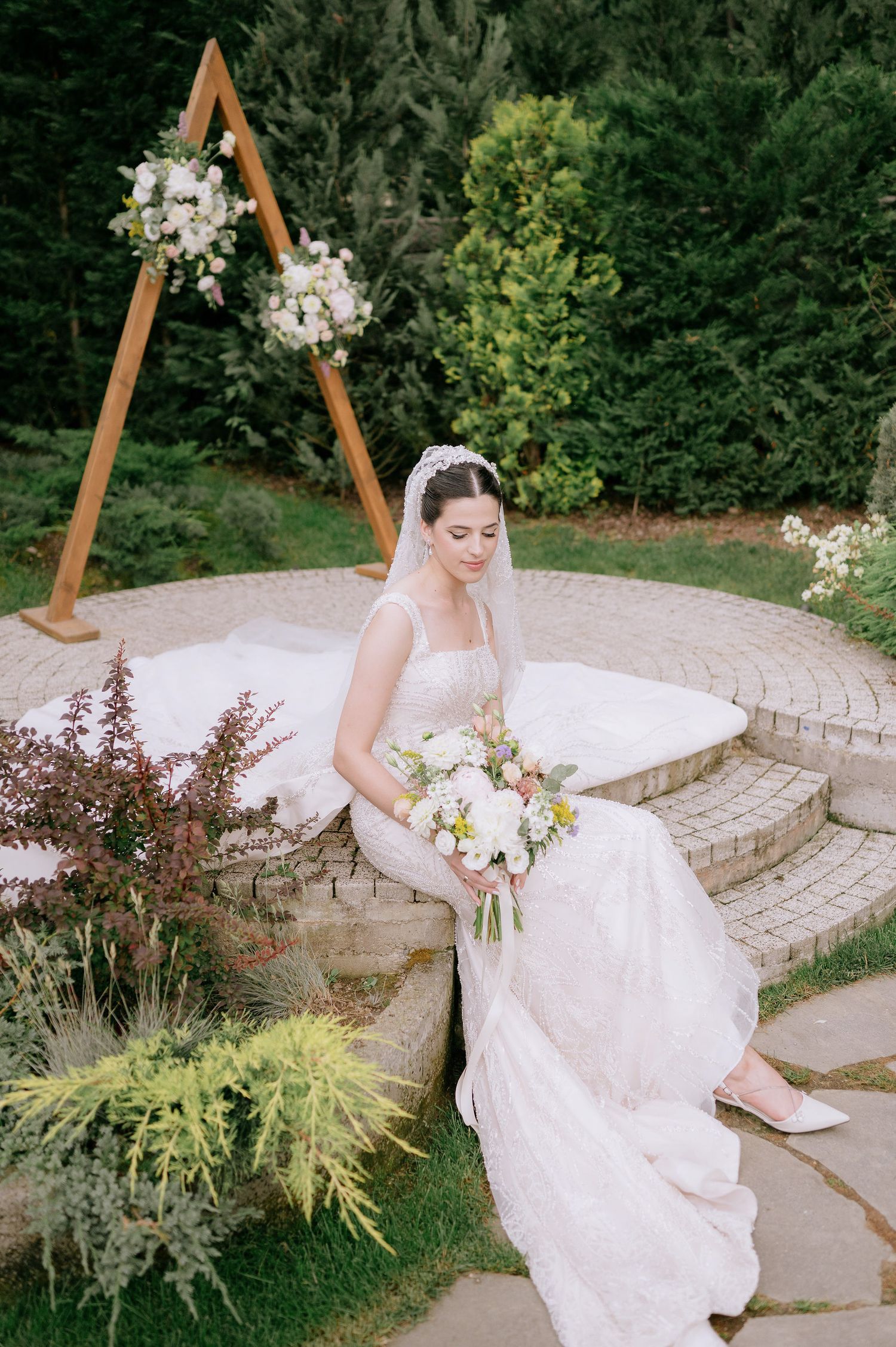 A bride in a white gown poses gracefully on stone steps surrounded by lush garden greenery.