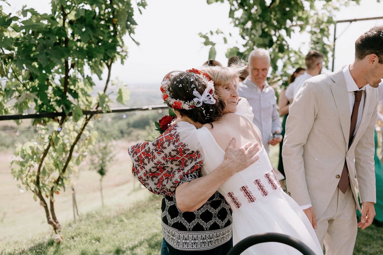 Wedding guests gather outdoors under green trees at a summer celebration.