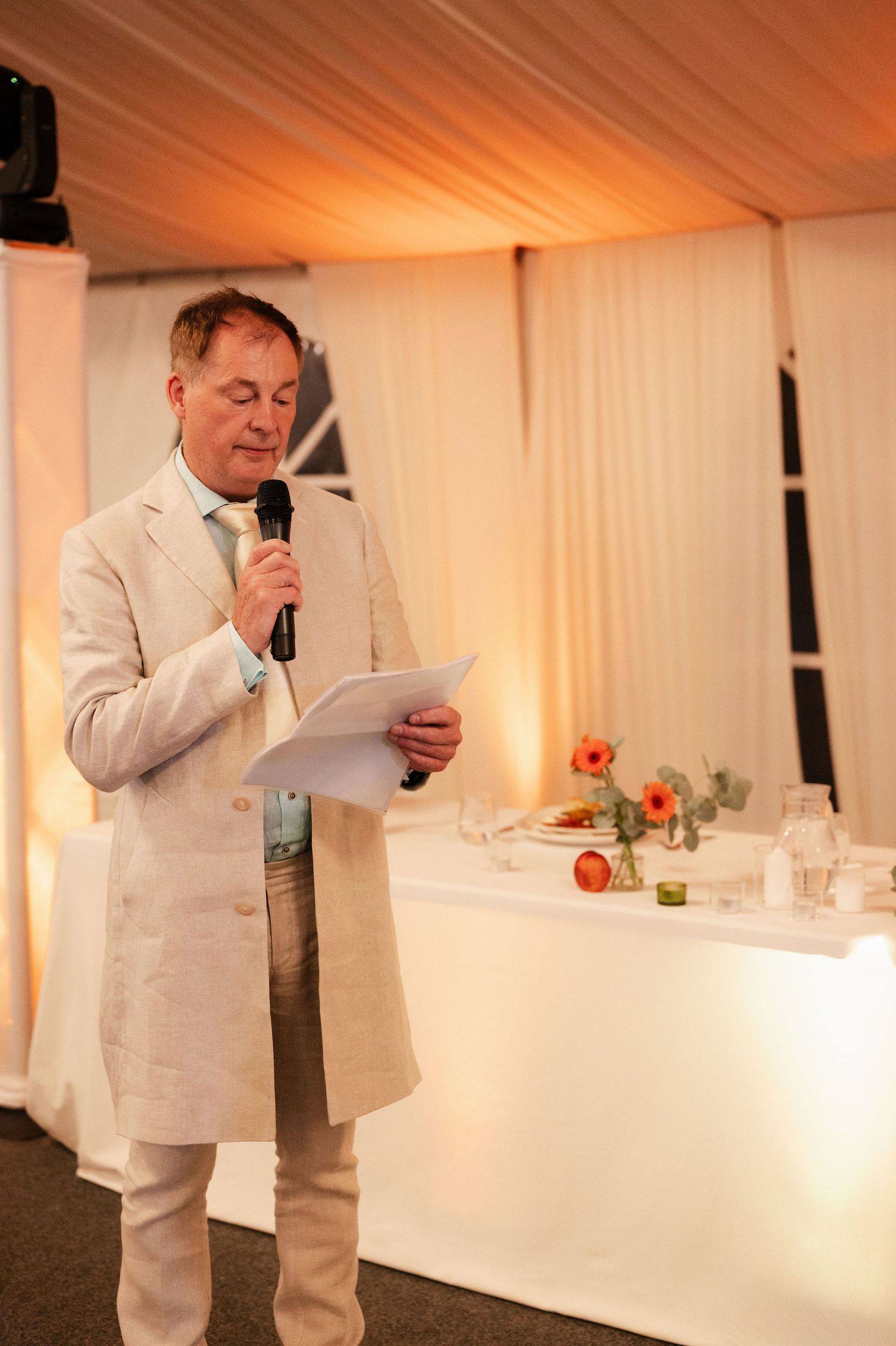 A speaker in a light-colored suit delivers remarks at an elegant reception while holding papers.