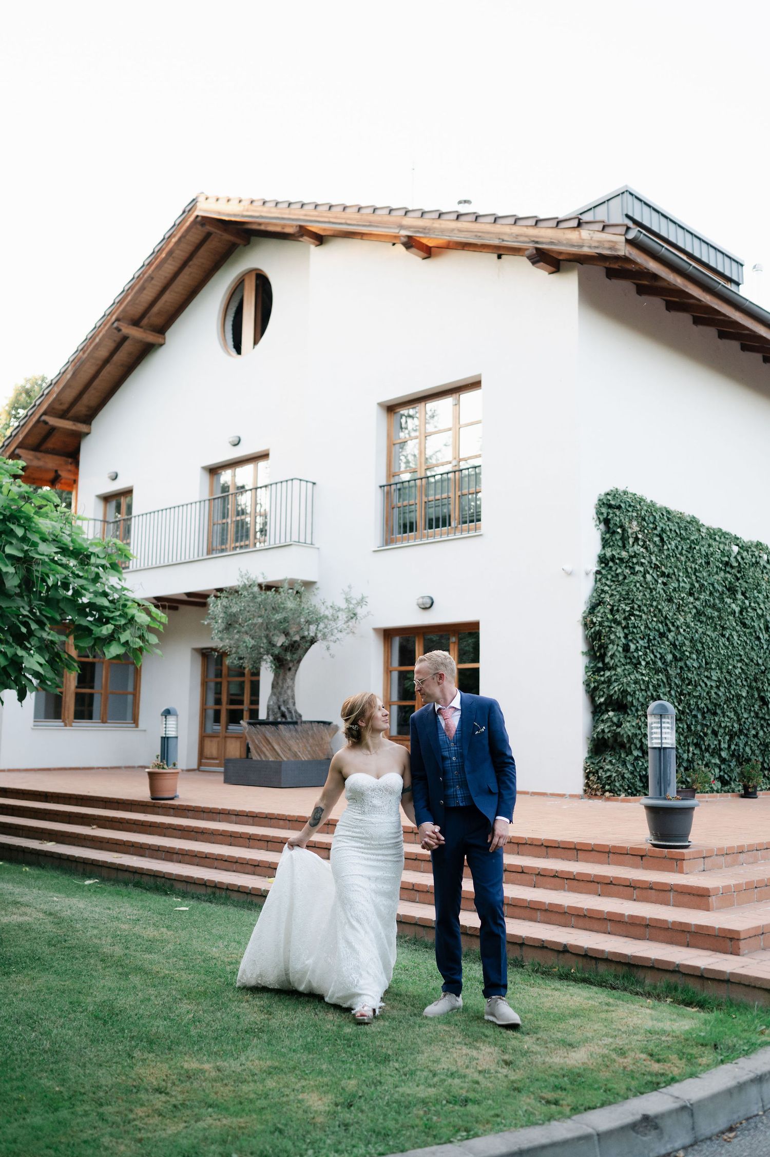 A wedding couple shares a tender moment on the lawn of an elegant white house with terrace steps.