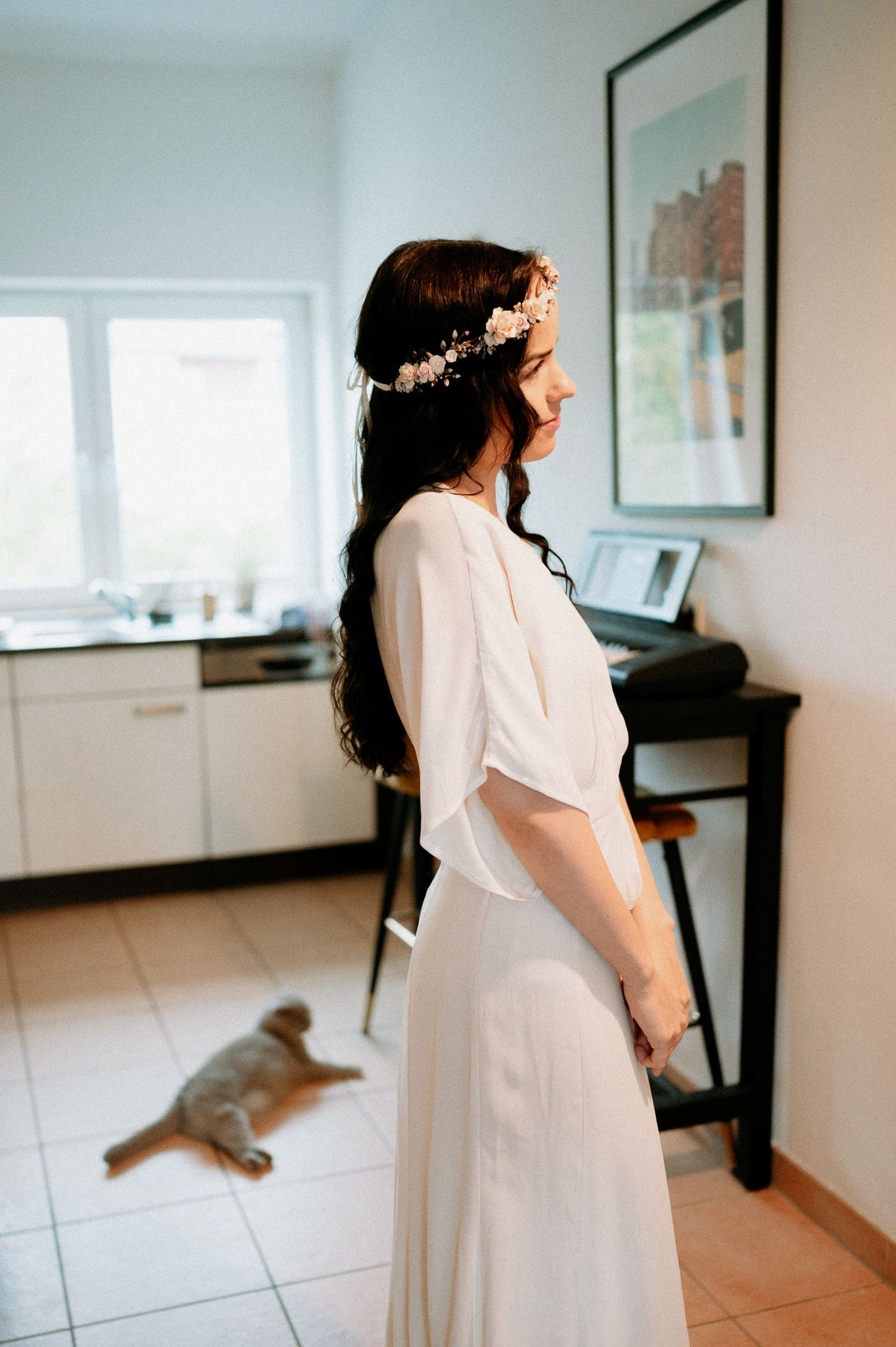 A person in a white dress stands in a bright modern room with a piano visible in the background.