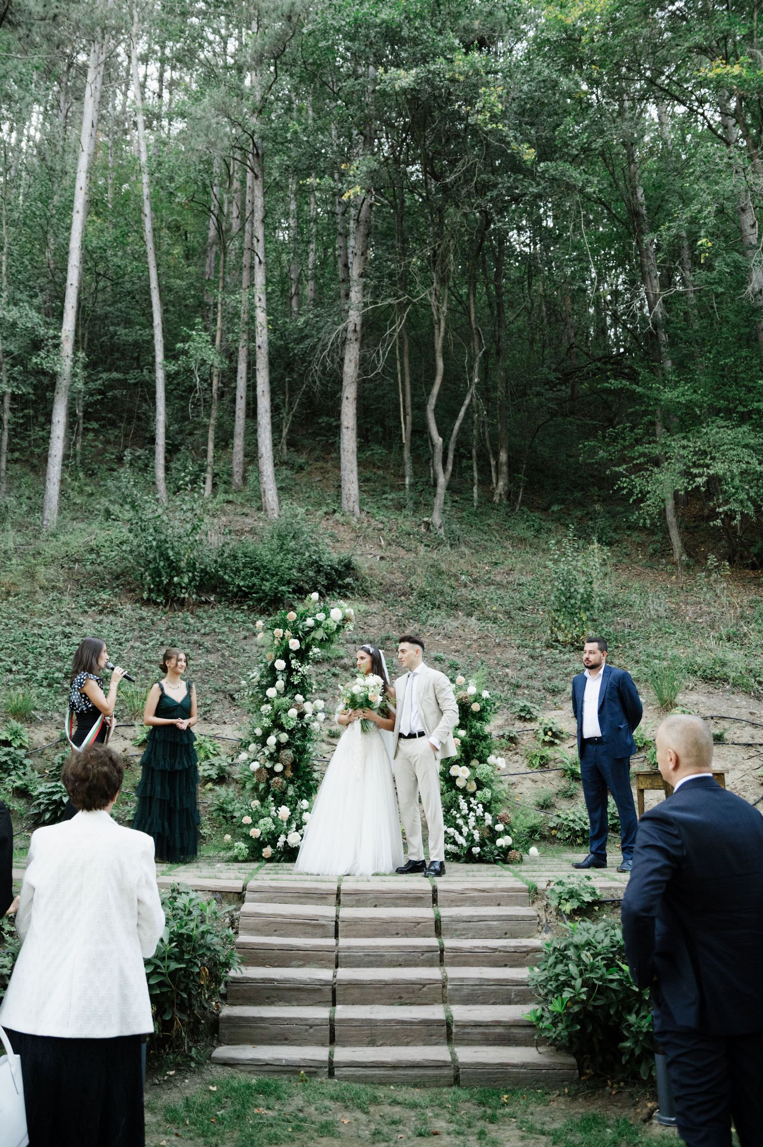 Forest wedding ceremony with guests watching from stone steps surrounded by white floral arrangements.