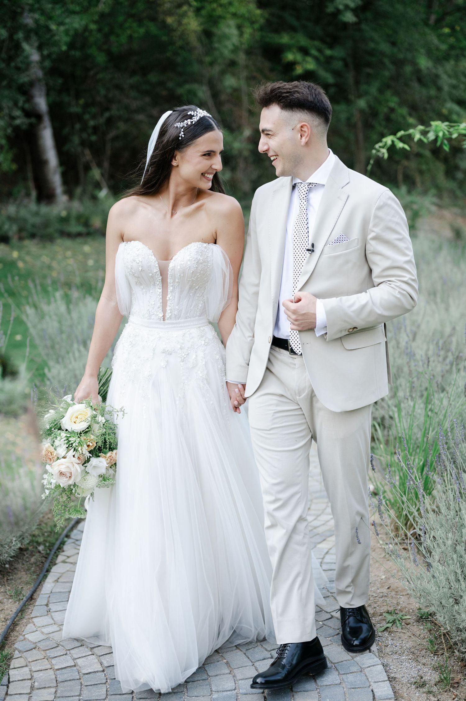 Wedding couple in white attire walking together through garden pathway.