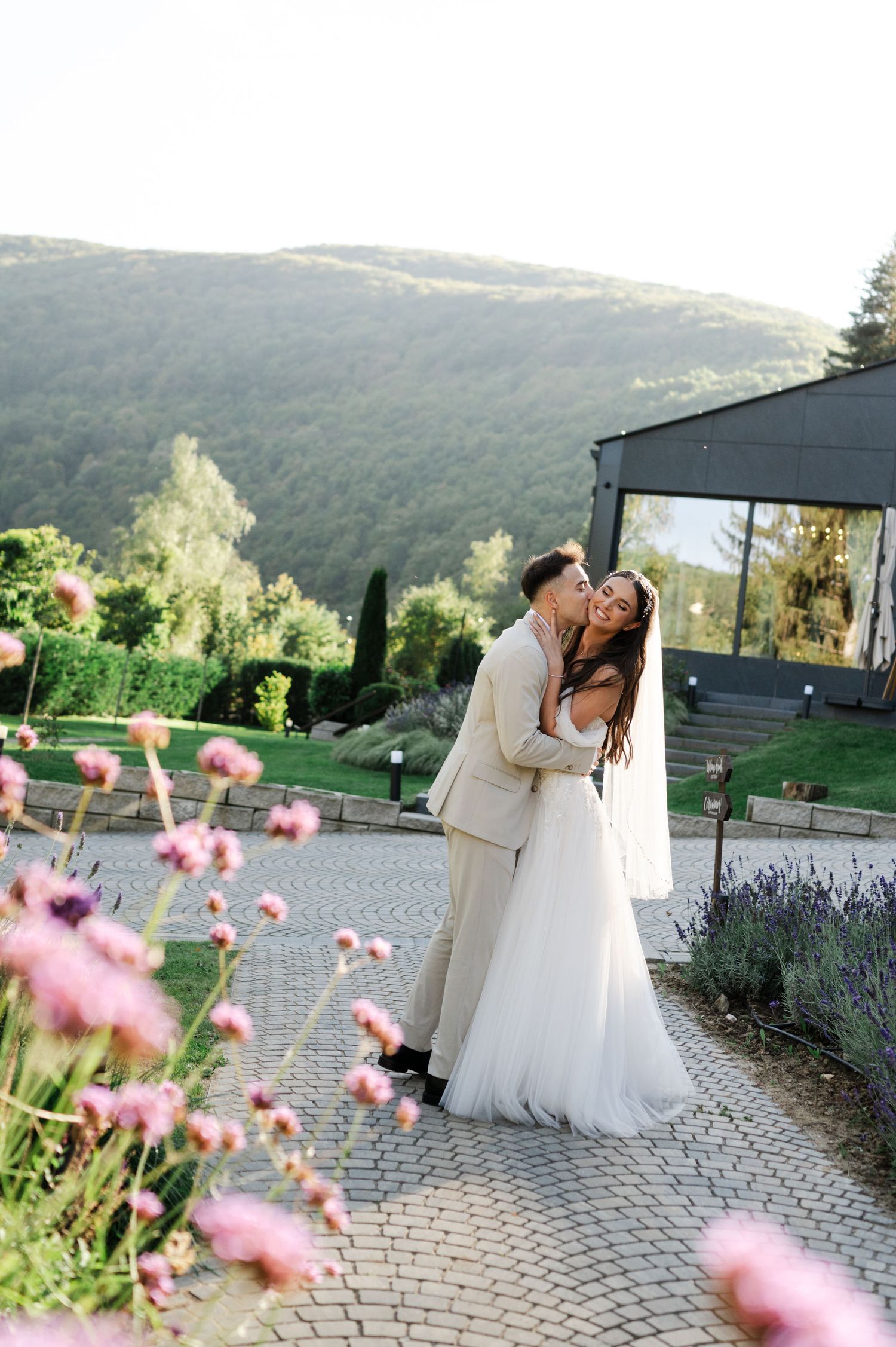 Wedding couple shares intimate moments on a stone path surrounded by pink flowers and mountain views.