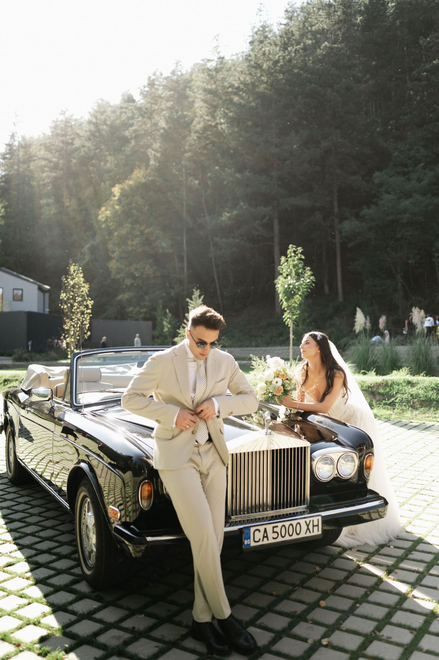 A couple in wedding attire poses playfully with a luxury Rolls Royce car outdoors.