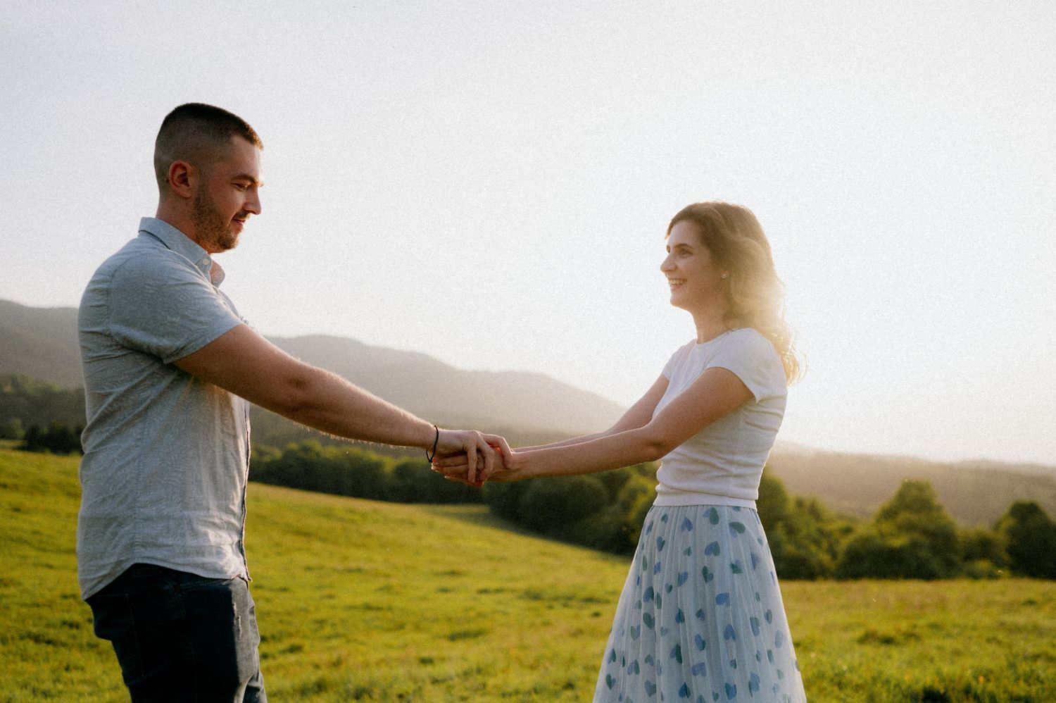A couple dances together in a sunlit green meadow with mountains in the background during golden hour.
