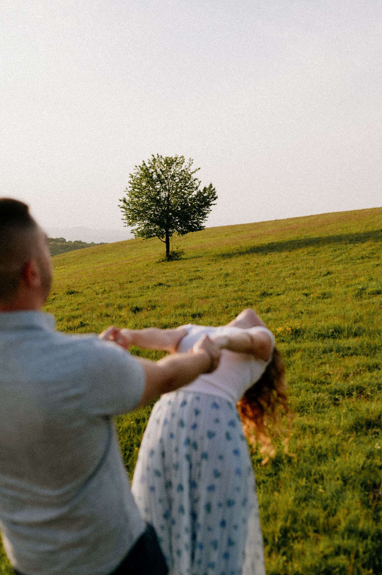 Dancing couple twirling in a grassy field with a lone tree on the hilltop at sunset.