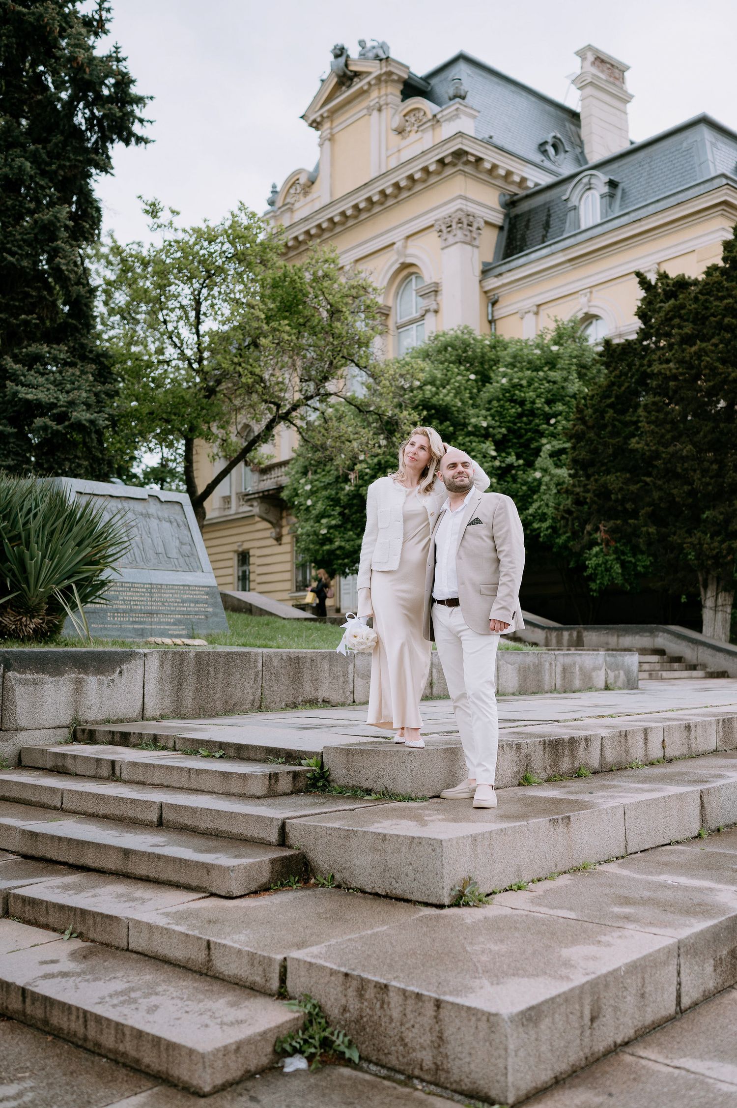 Couple dressed in all white poses on stone steps of a classical architecture building.
