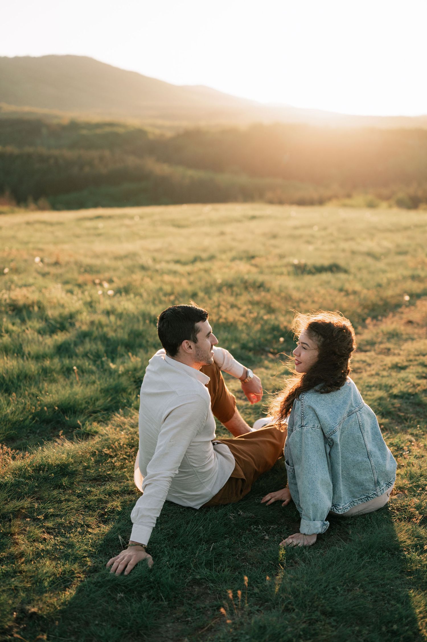 A couple lying on grassy hillside during golden sunset hour enjoying a peaceful moment together.