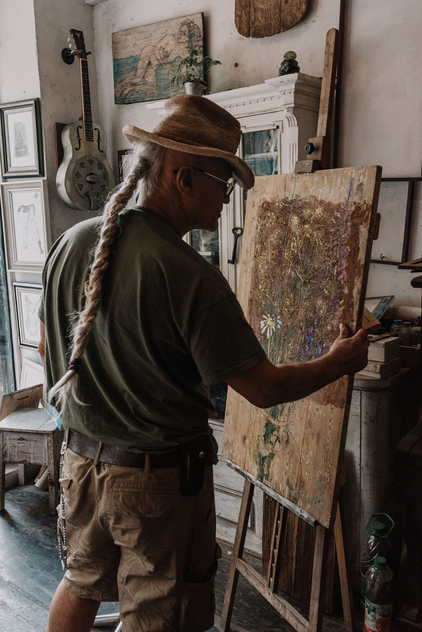 Artist in casual attire and hat working on a painting at an easel in a rustic studio setting.