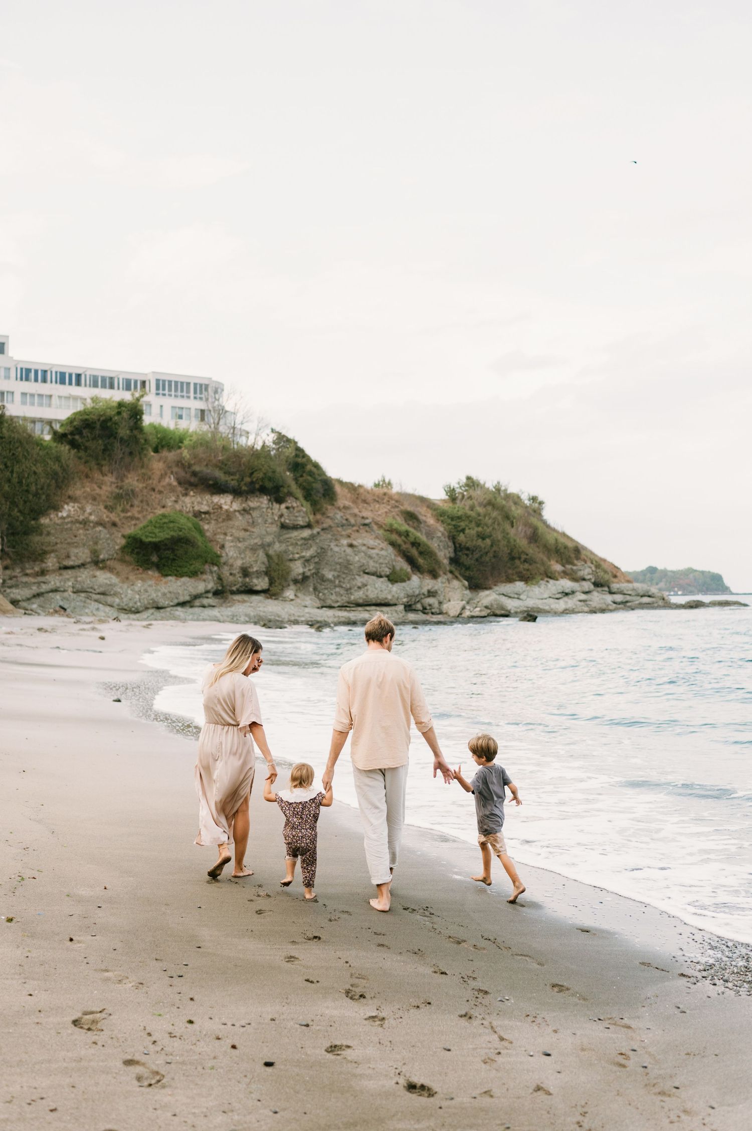 Family of four walking hand-in-hand along a scenic beach shoreline with rocky cliffs and a white building in the background.
