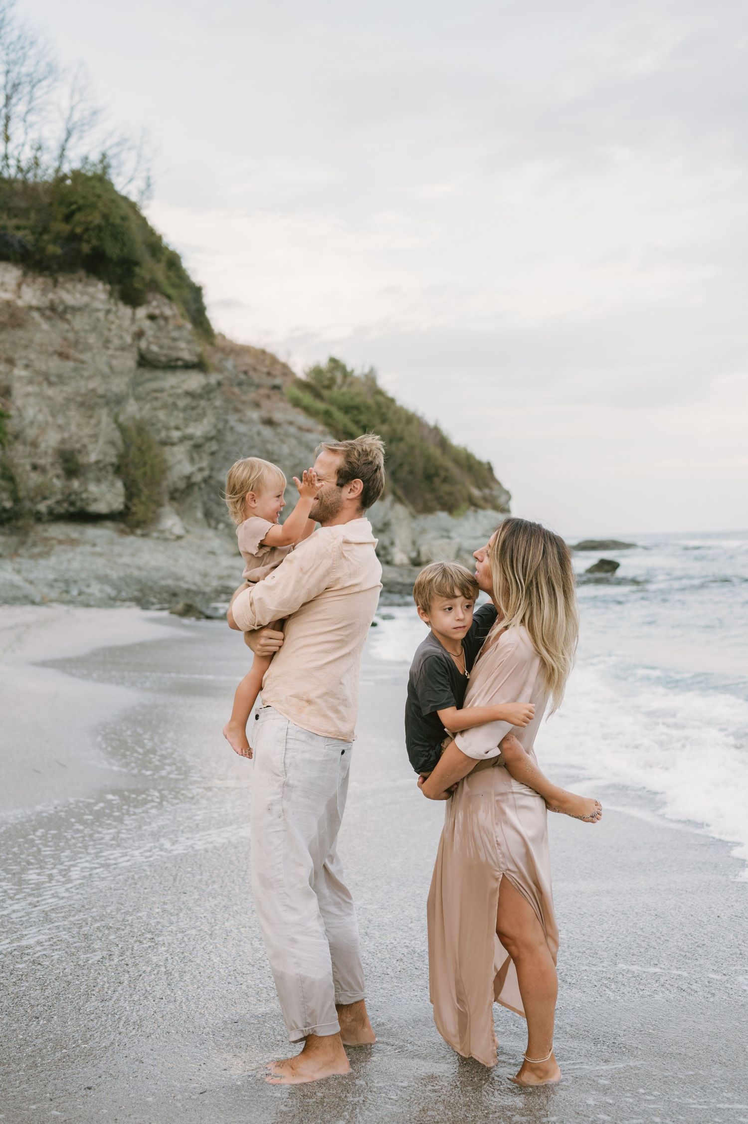 Family of four enjoying a beach day, parents holding young children near ocean waves with rocky cliffs in background.