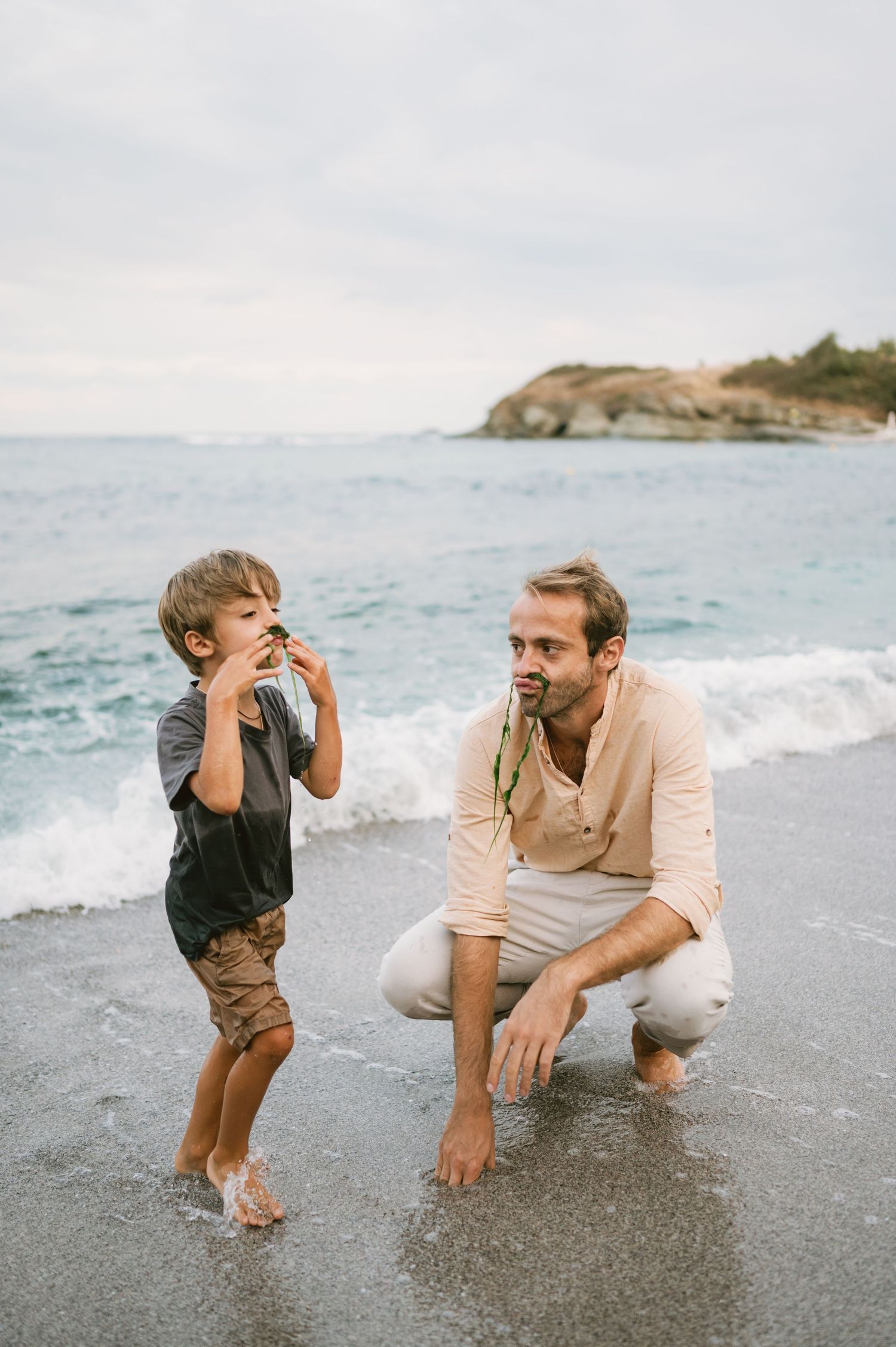 A father crouches on a sandy beach while his young son shows him a shell, with ocean waves in the background.