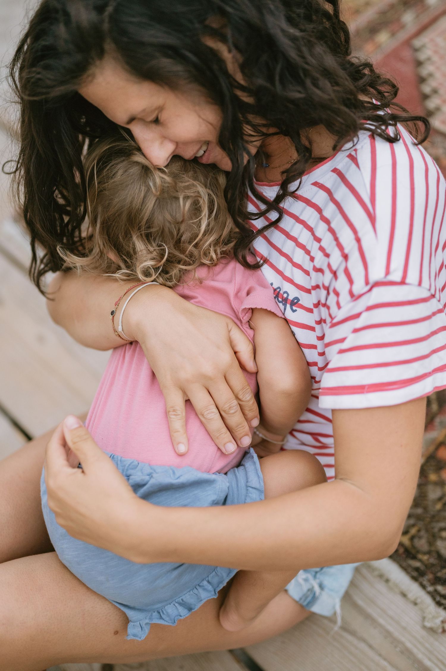 A mother with curly dark hair hugs her young toddler girl tenderly outdoors, both wearing pink and blue outfits.