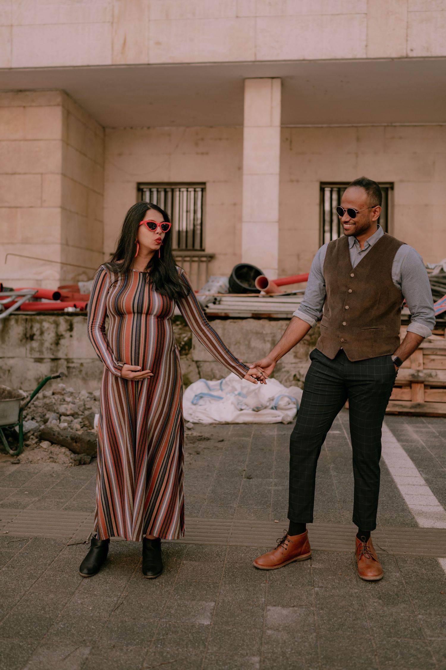 Pregnant couple holding hands outdoors, woman in striped maxi dress, man in vest and trousers, both wearing sunglasses.