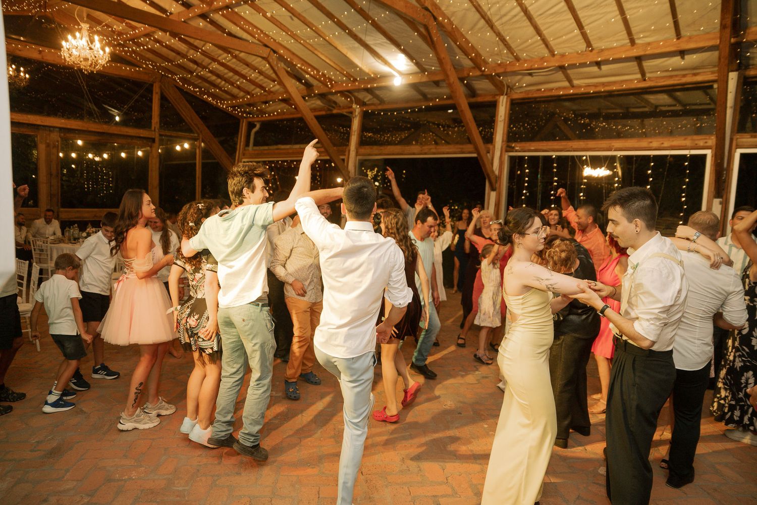 Wedding guests dance and celebrate on a wooden dance floor inside a rustic barn venue lit by string lights and chandeliers.