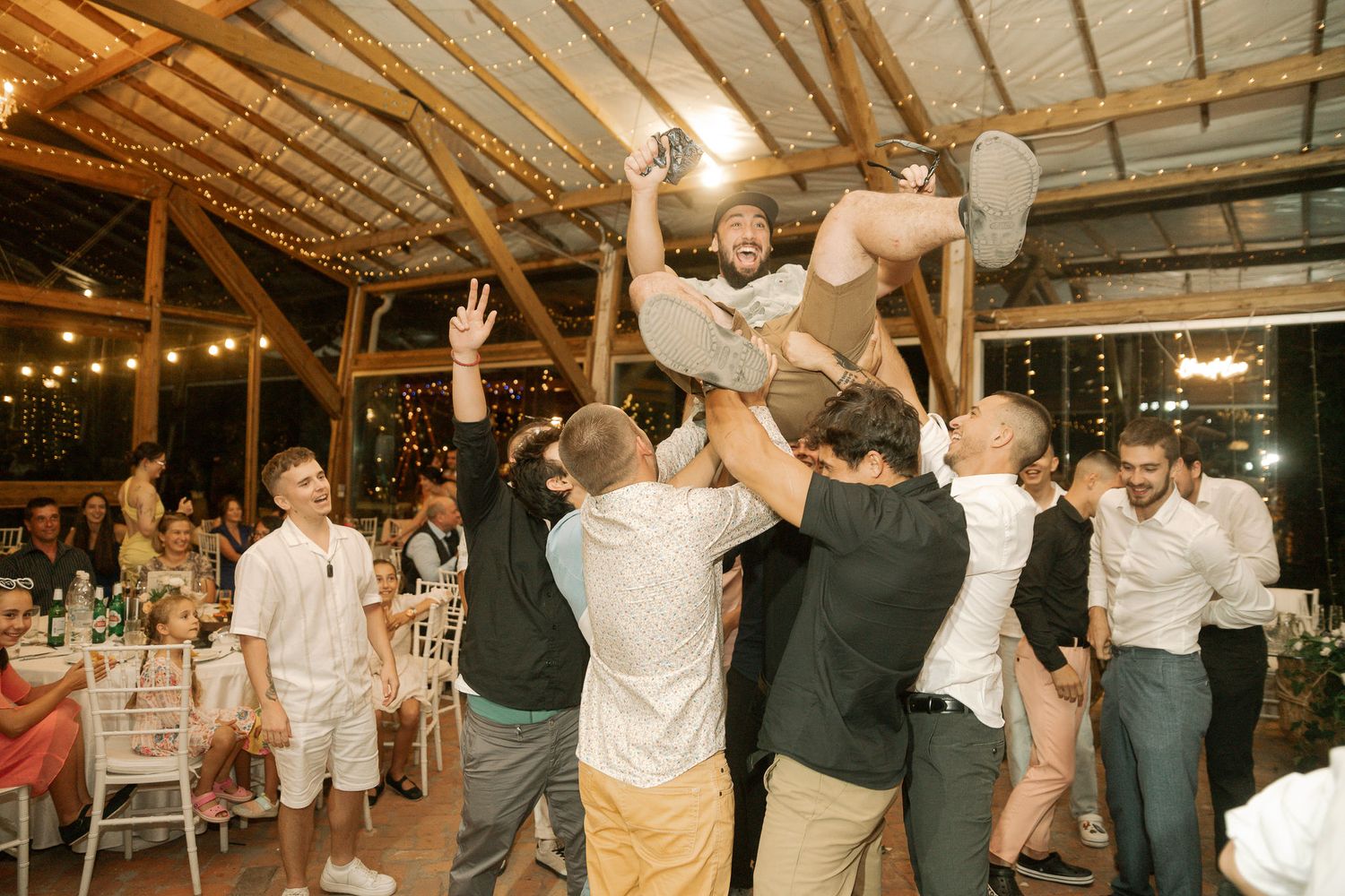 Wedding guests dance together in a rustic barn venue with wooden beams and twinkling lights overhead.