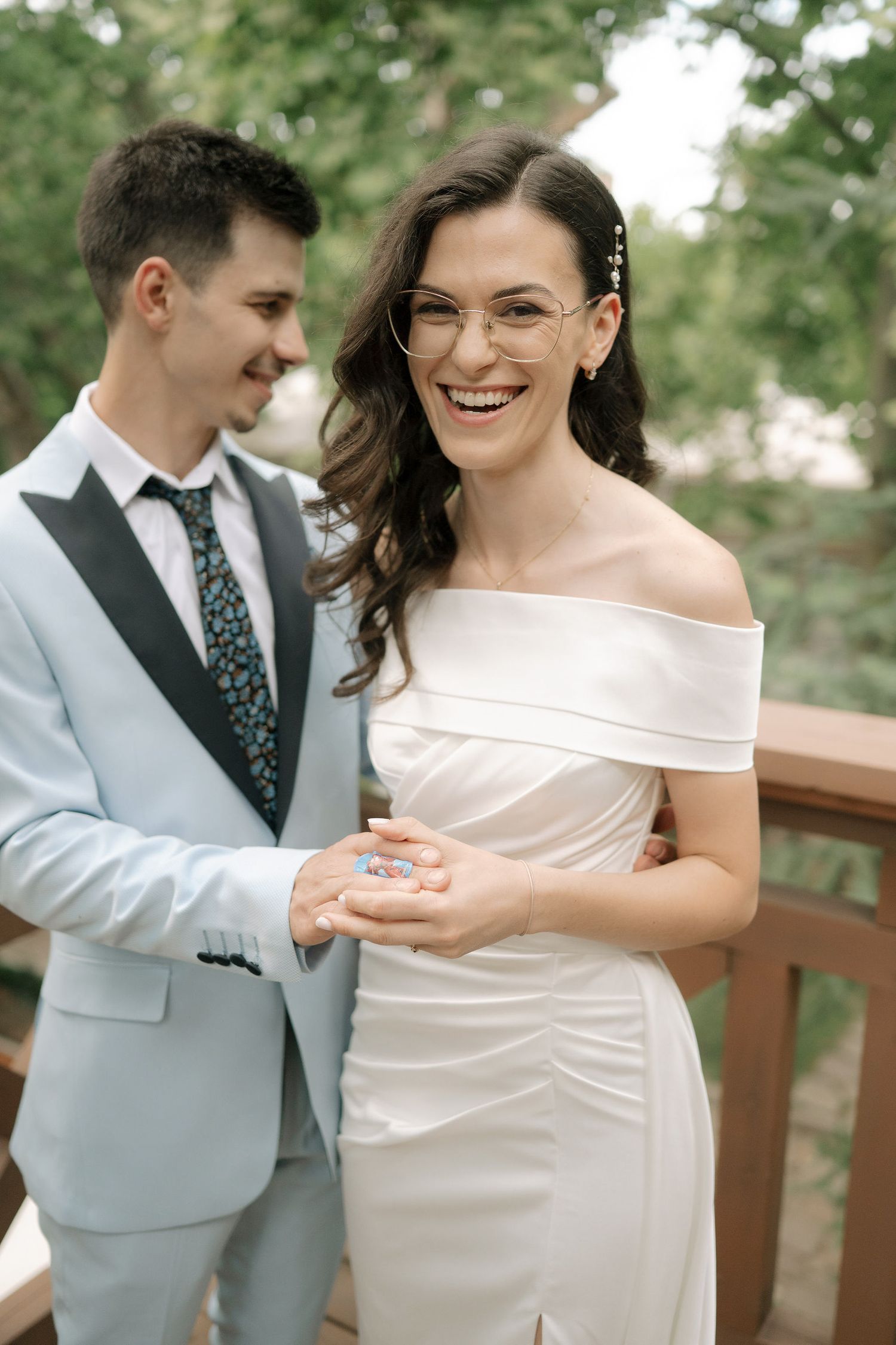 A couple shares a joyful moment on a wooden deck, with the bride wearing an elegant off-shoulder white dress.