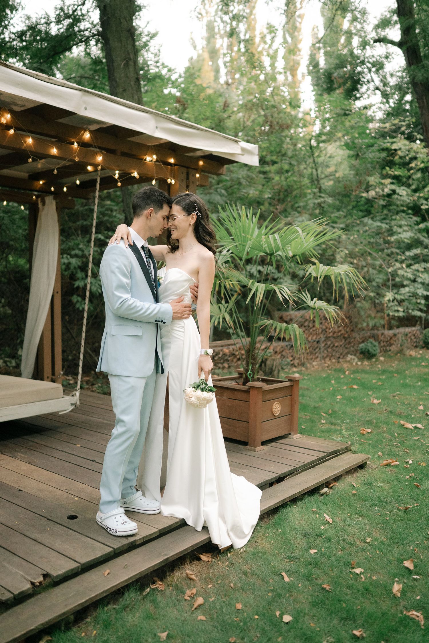 Couple shares romantic moments on a wooden deck surrounded by lush greenery and string lights at an outdoor wedding venue.