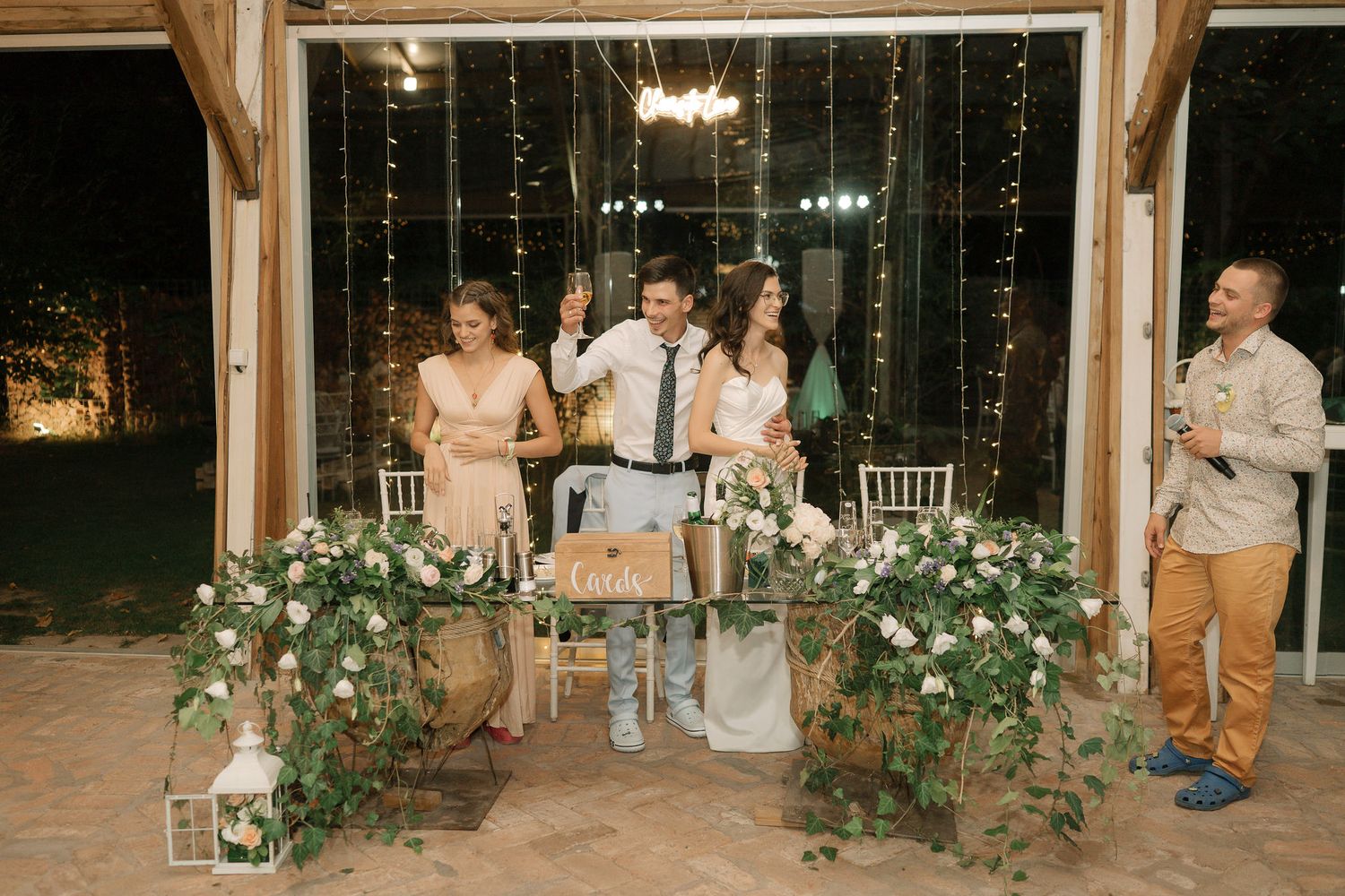 Wedding party gathers in a rustic venue decorated with greenery and lanterns at night.