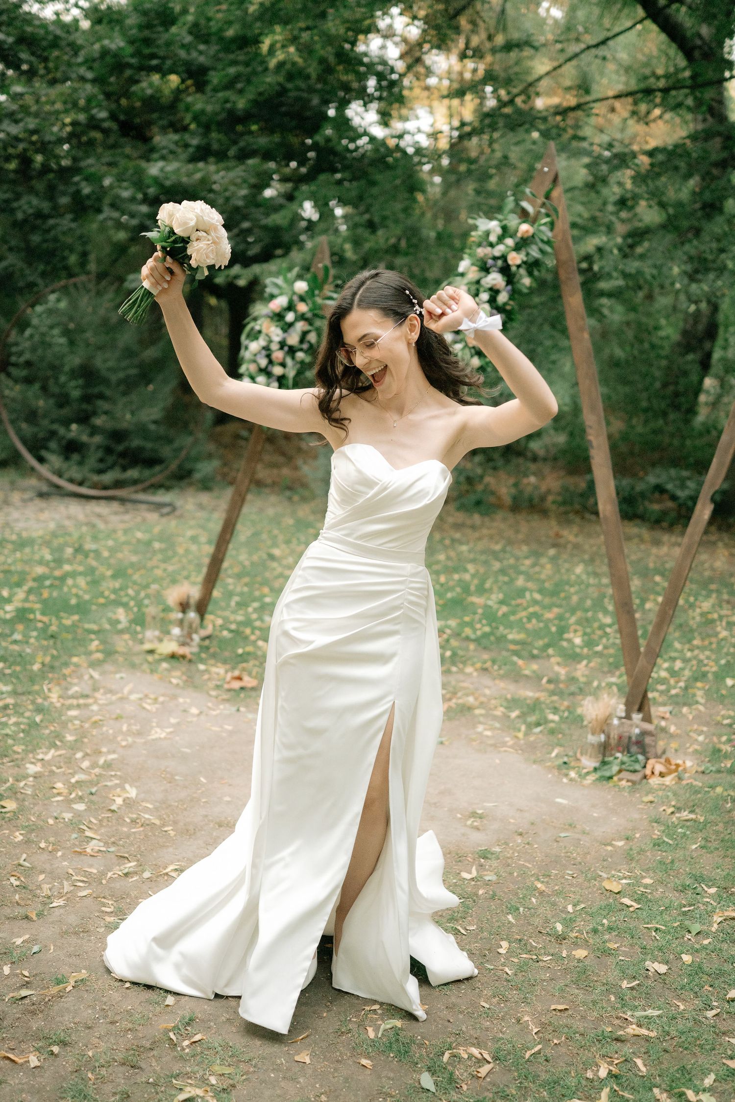 A joyful bride in a white wedding dress dances and twirls through a wooded outdoor setting during a photo session.