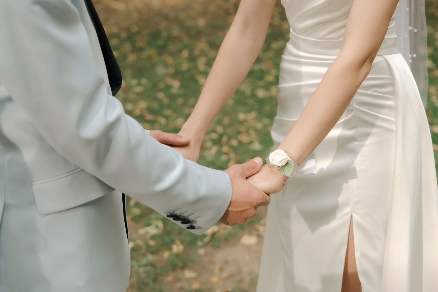 A couple in wedding attire holds hands while walking together in a garden setting with fallen autumn leaves.