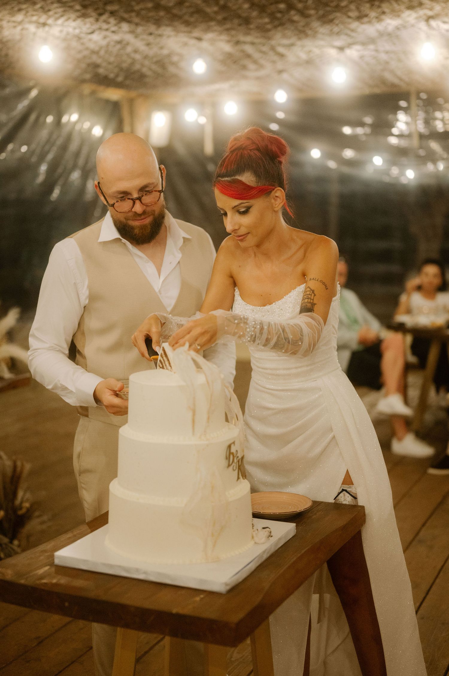Couple cutting into their elegant white wedding cake together during an evening reception with string lights overhead.