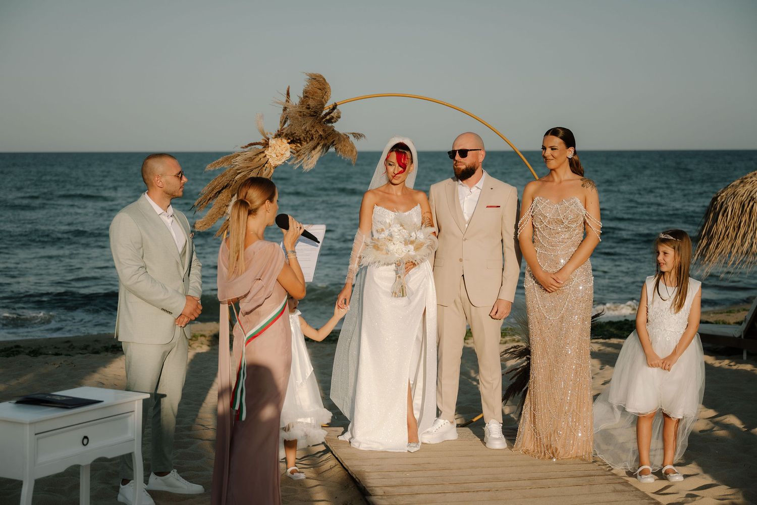 A beachside wedding ceremony takes place on a wooden deck with an arched palm frond backdrop against the ocean.