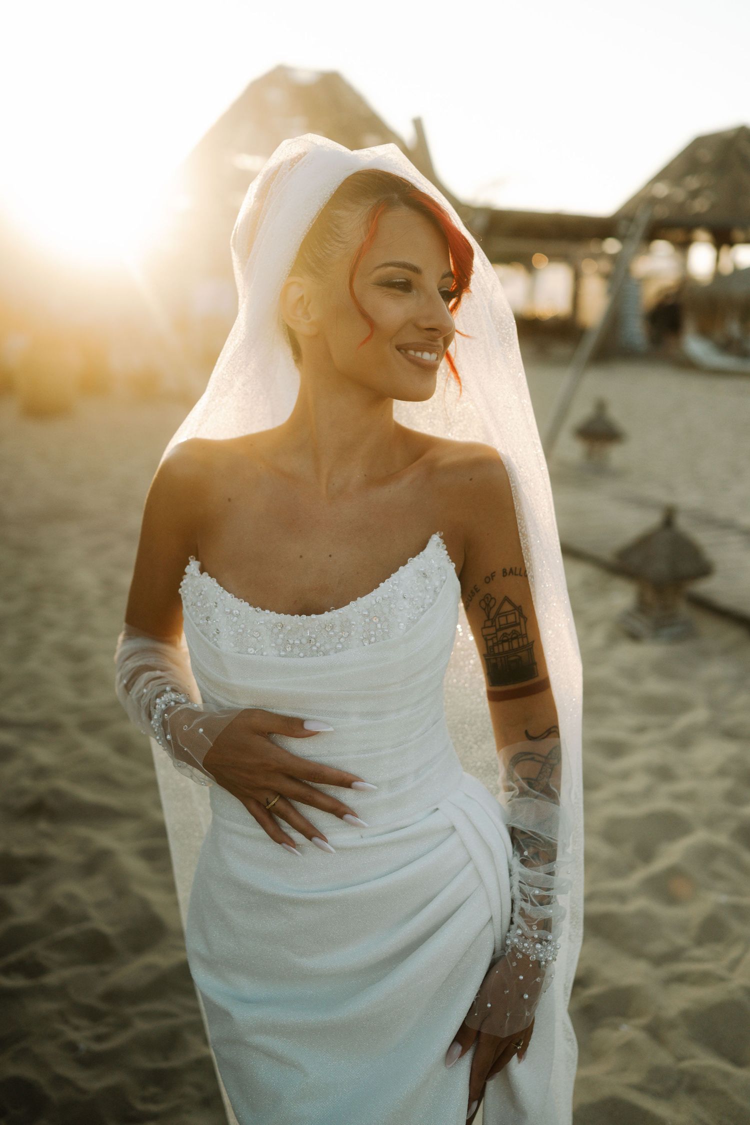 A person in a white wedding dress poses on a sandy beach at sunset while showing off detailed arm tattoos.