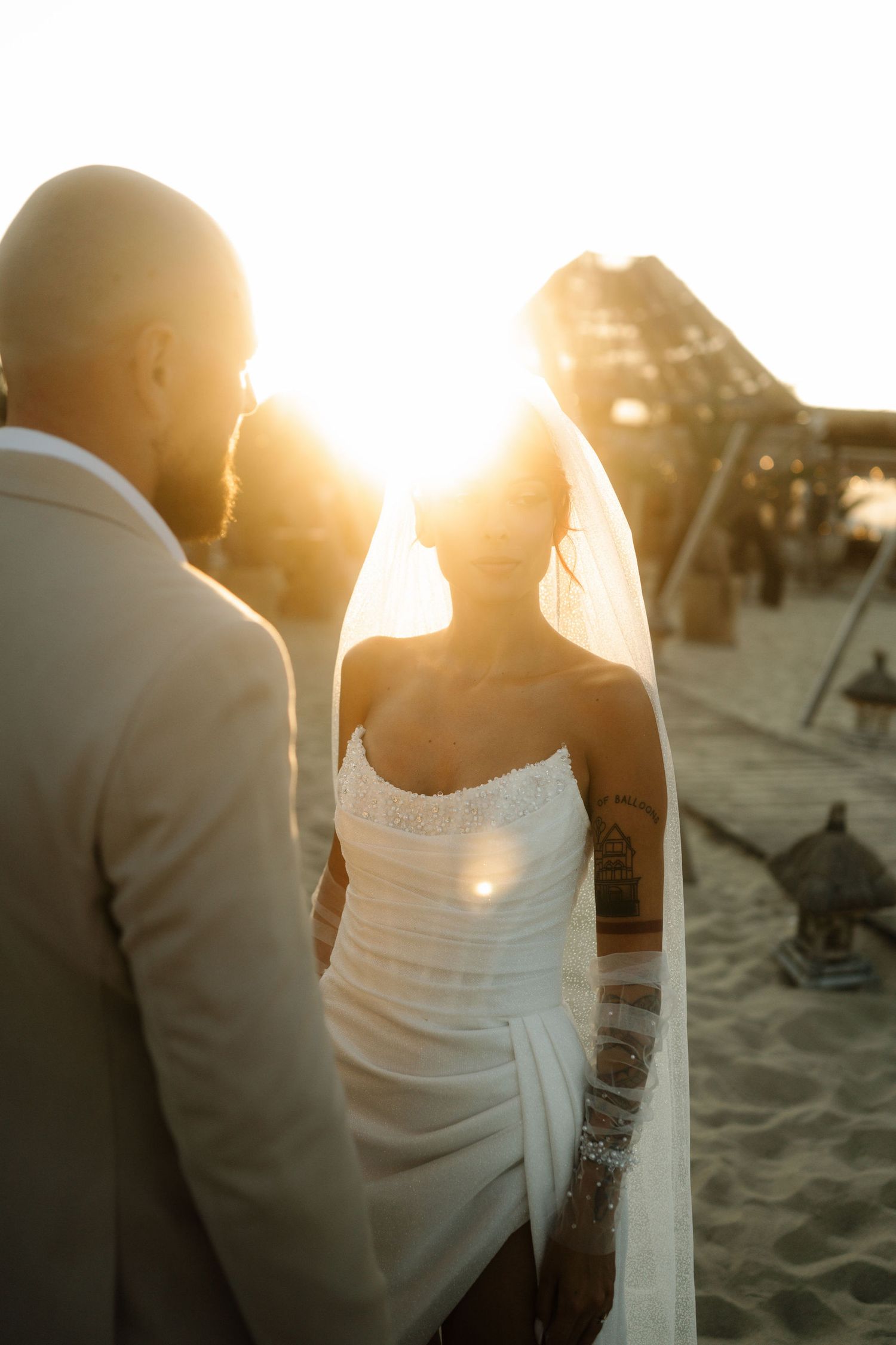 A romantic beach scene at sunset with silhouetted figures sitting on the sand as golden light streams through.