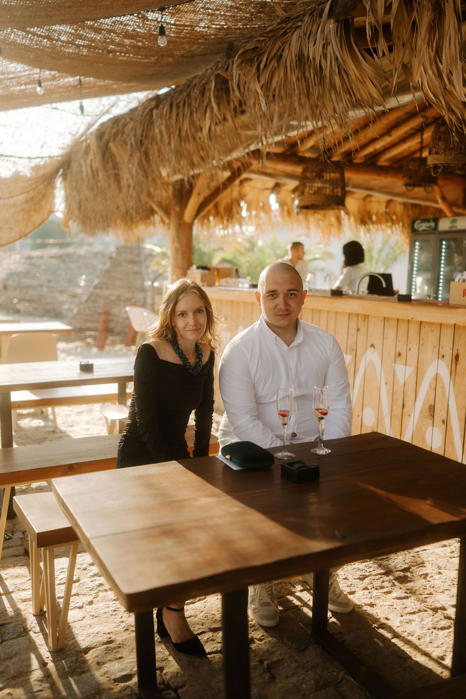 Couple sits together at rustic wooden tables under a thatched roof patio at a beachside restaurant.
