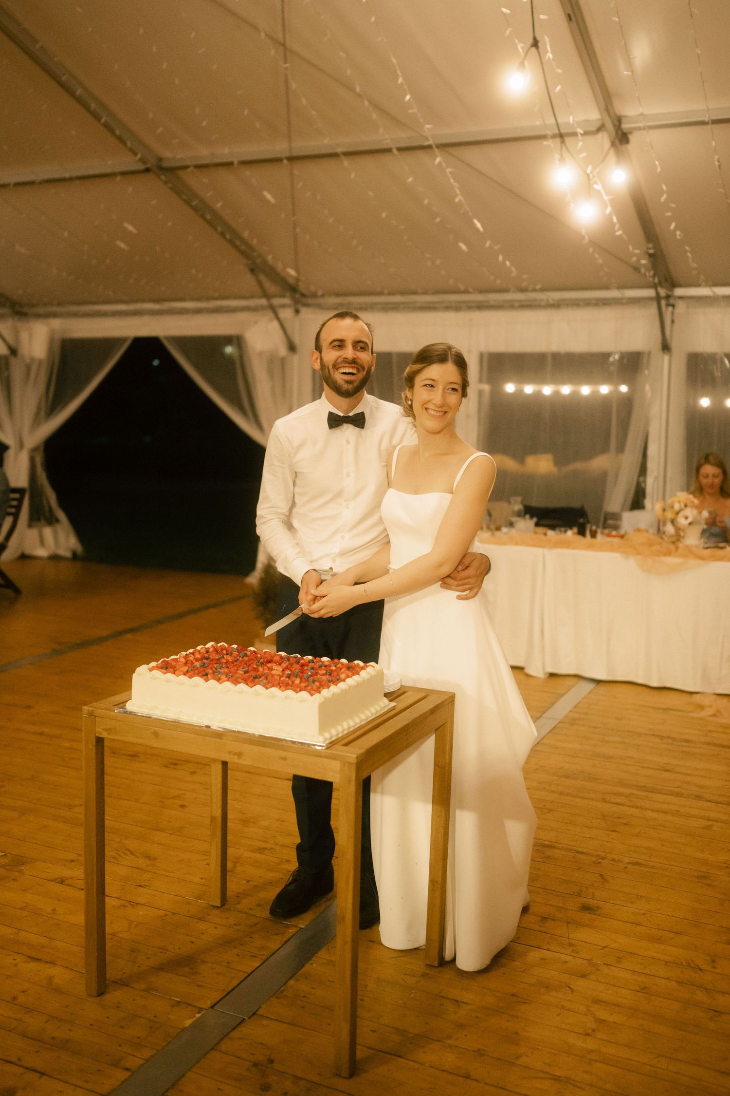 Two people in formal attire carry a large white wedding cake across a wooden floor in an illuminated reception tent.