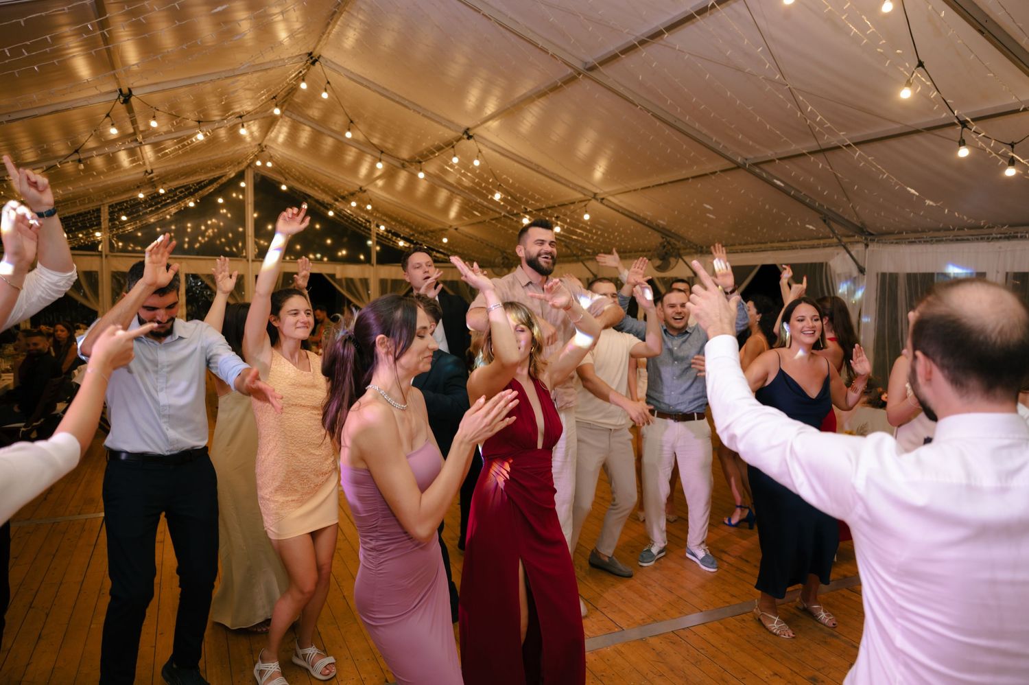 Wedding guests dancing and celebrating on a dance floor inside a reception venue with warm lighting.