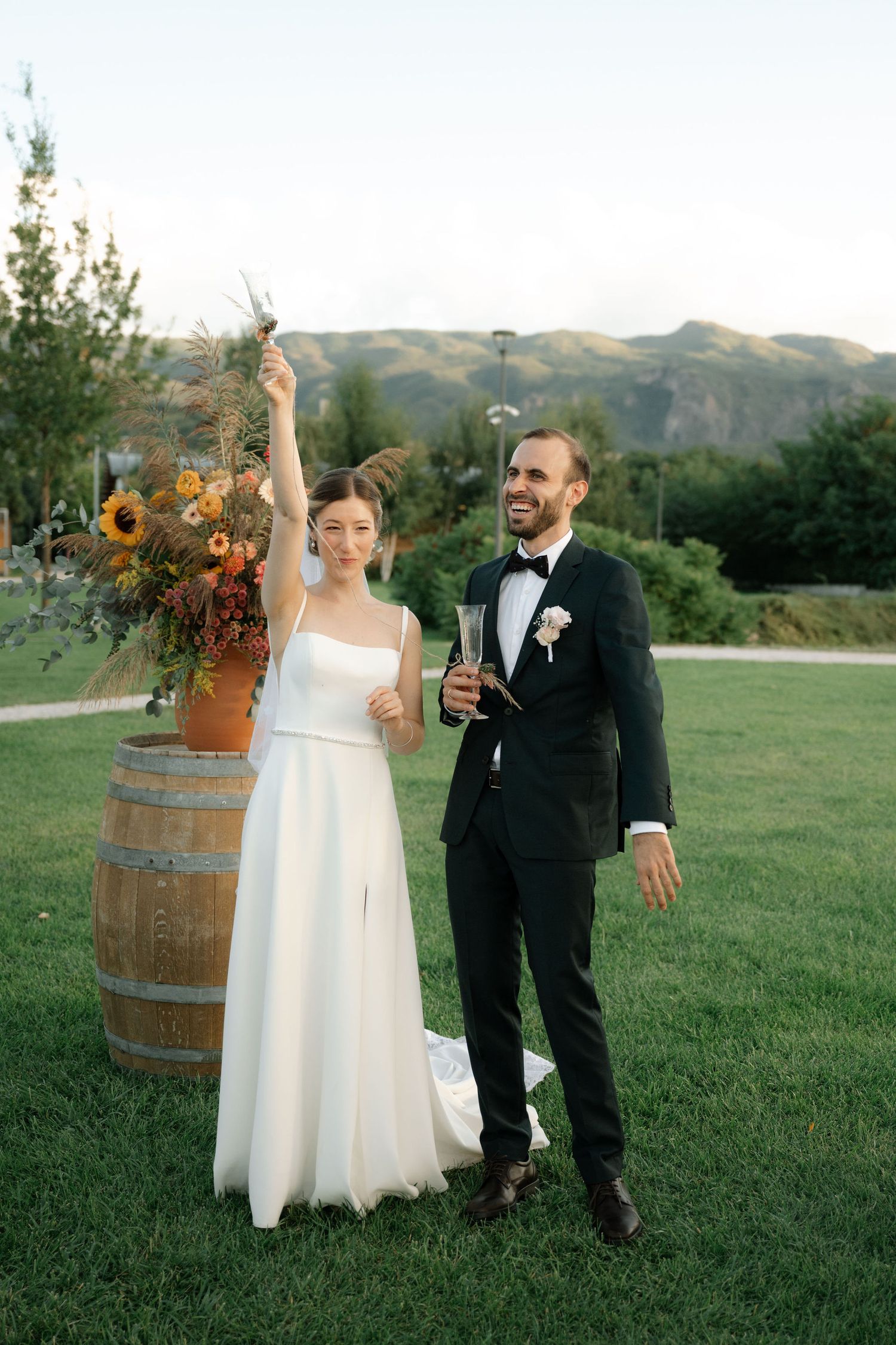 A couple celebrates during their outdoor wedding reception next to rustic wine barrels at sunset in a grassy field.