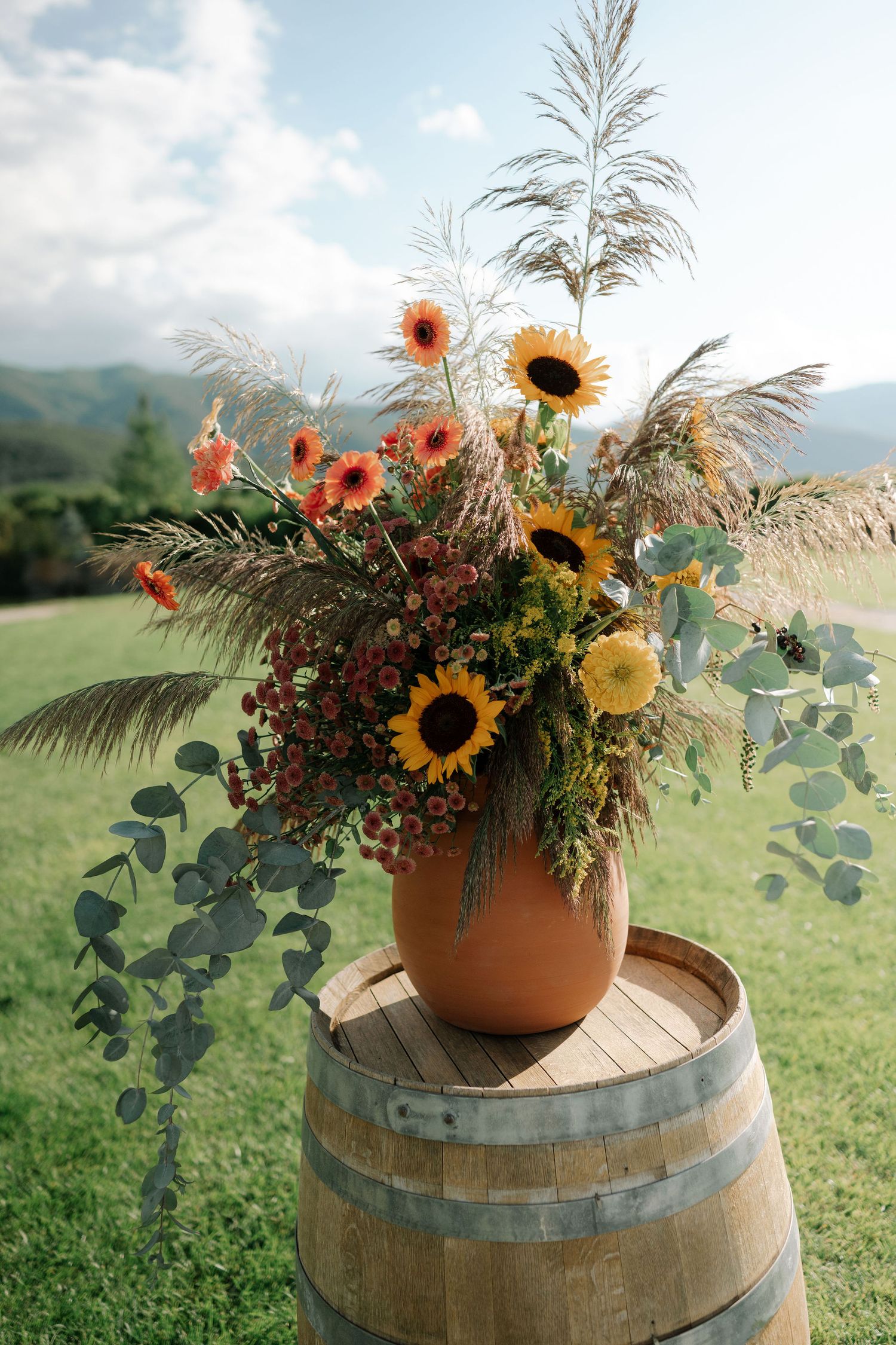 Rustic floral arrangement with sunflowers and wildflowers in terra cotta pot displayed on wooden wine barrel outdoors.