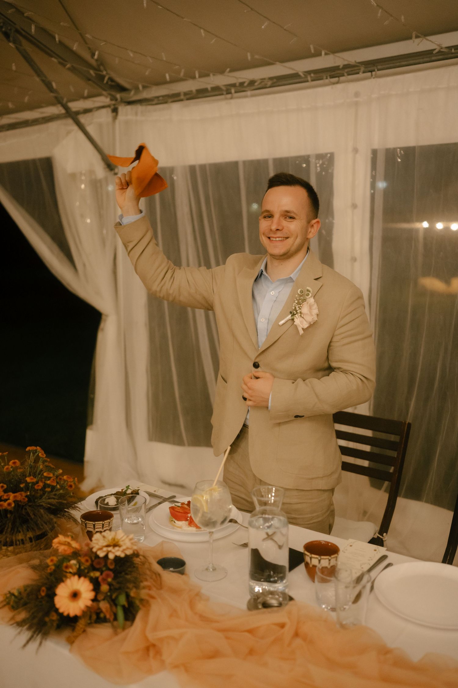 Wedding reception dinner table settings decorated with orange floral arrangements and warm lighting under a tent.