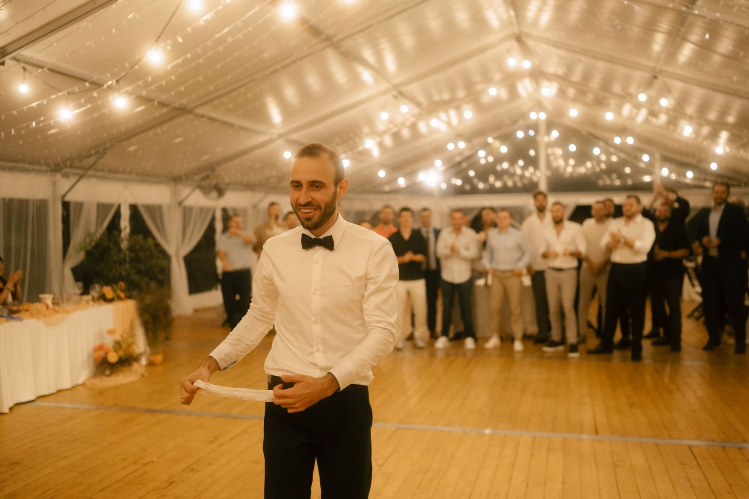Wedding celebration under twinkling string lights with guests gathered on a wooden dance floor.