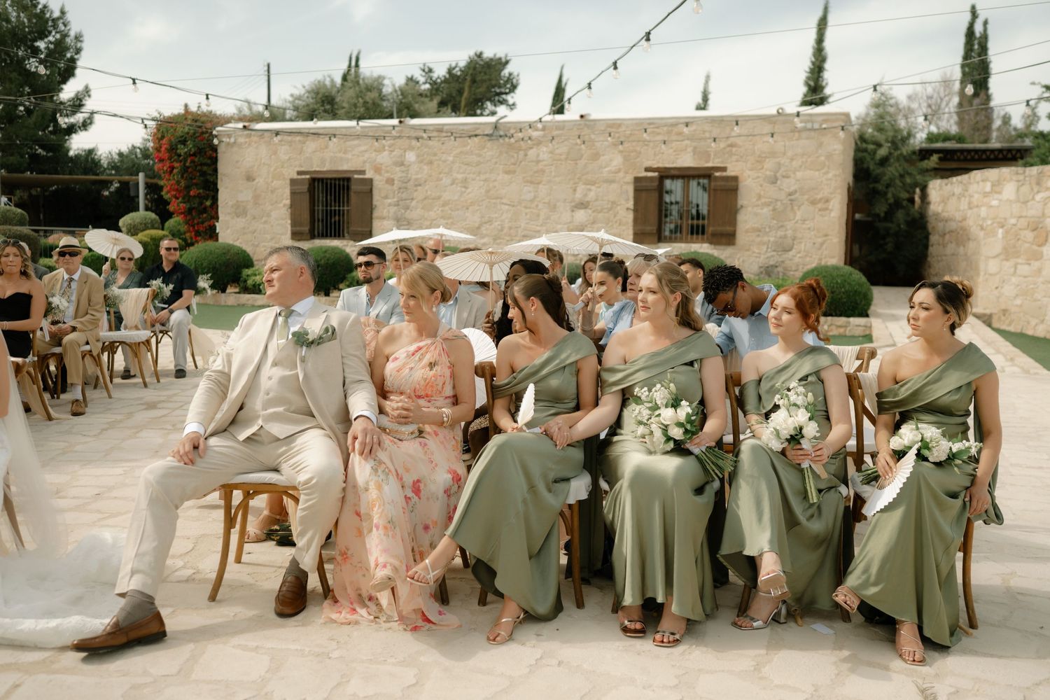 Wedding guests in sage green bridesmaid dresses and formal attire seated outdoors at a rustic stone venue ceremony.