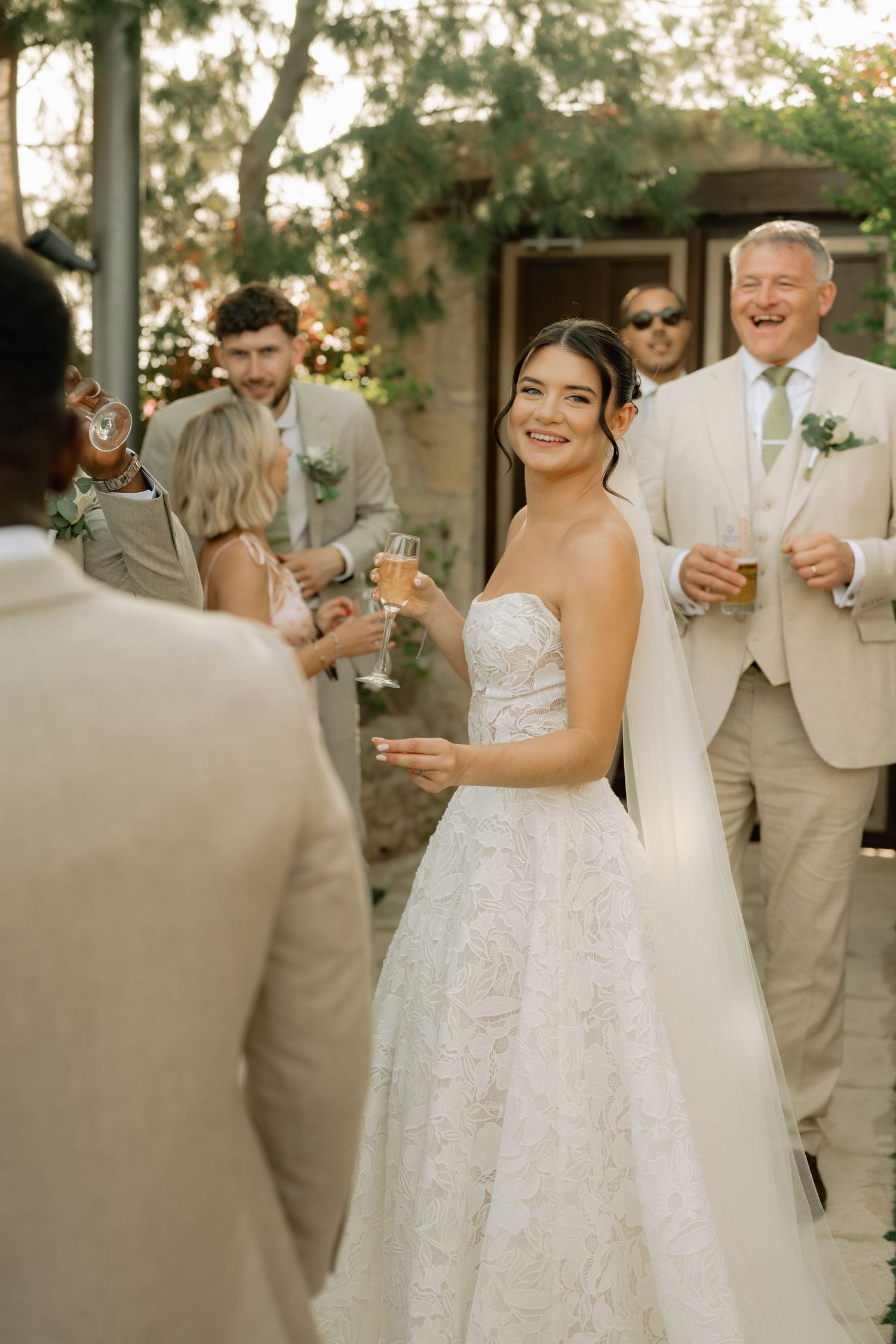 Smiling bride in strapless lace gown holds champagne surrounded by wedding guests outdoors.