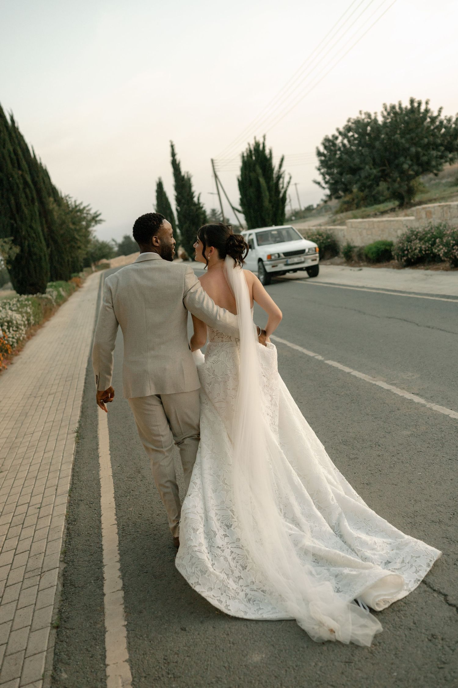 Bride and groom walking hand in hand on a road, bride in lace gown with long train, groom in beige suit.