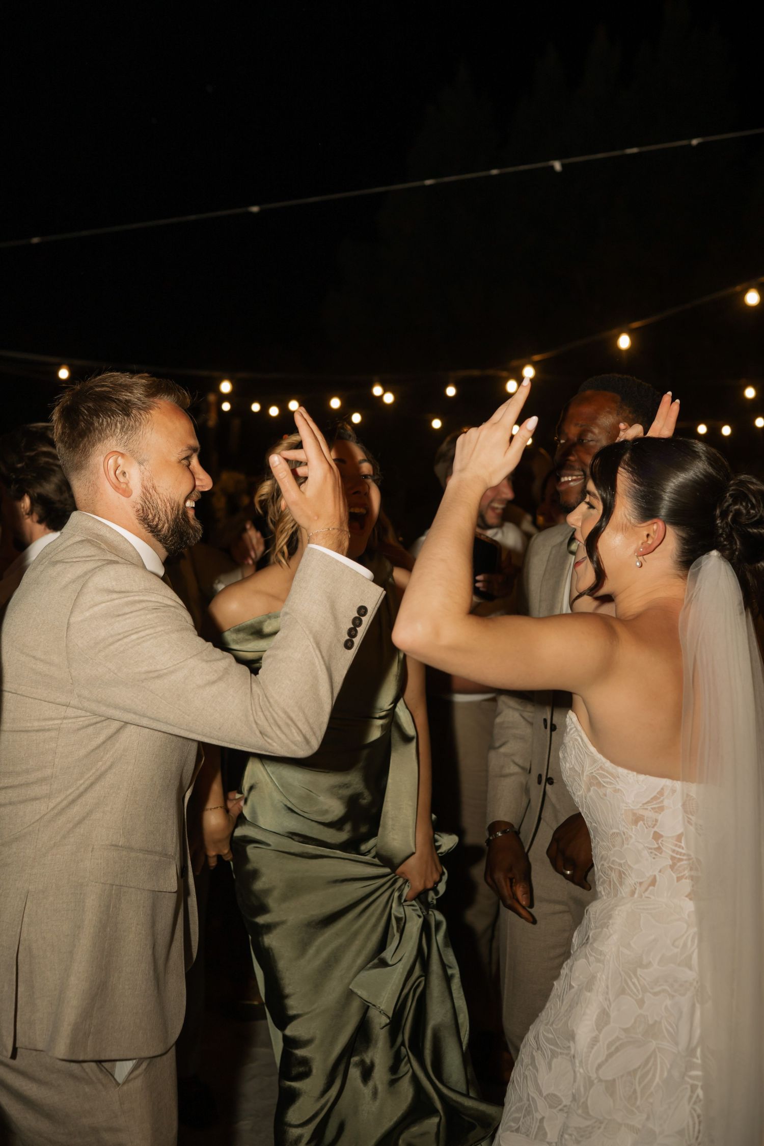 Bride and groom dancing joyfully at outdoor wedding reception under string lights surrounded by guests at night.