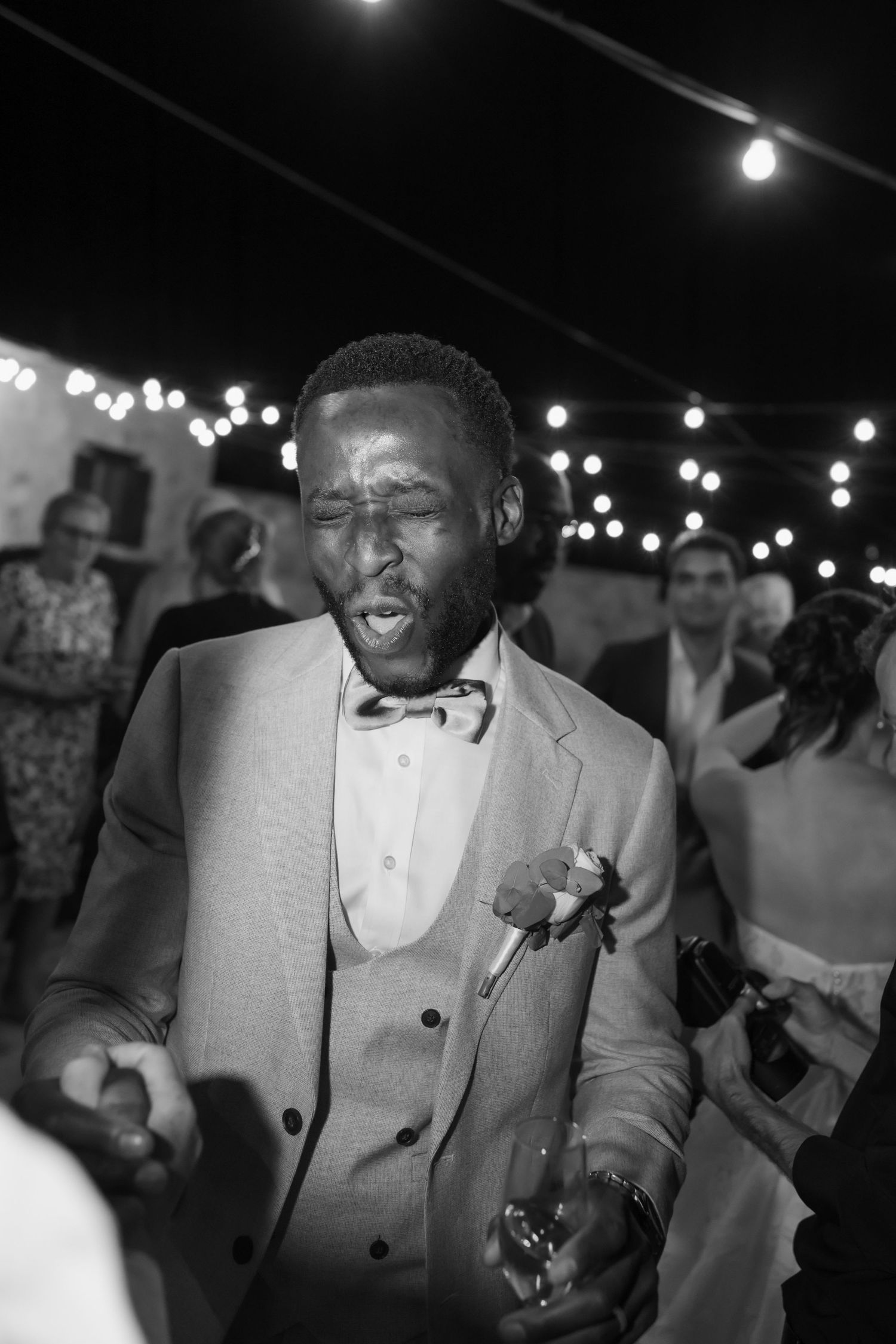 Man in suit and bow tie dancing joyfully at a wedding reception under string lights, black and white photo.