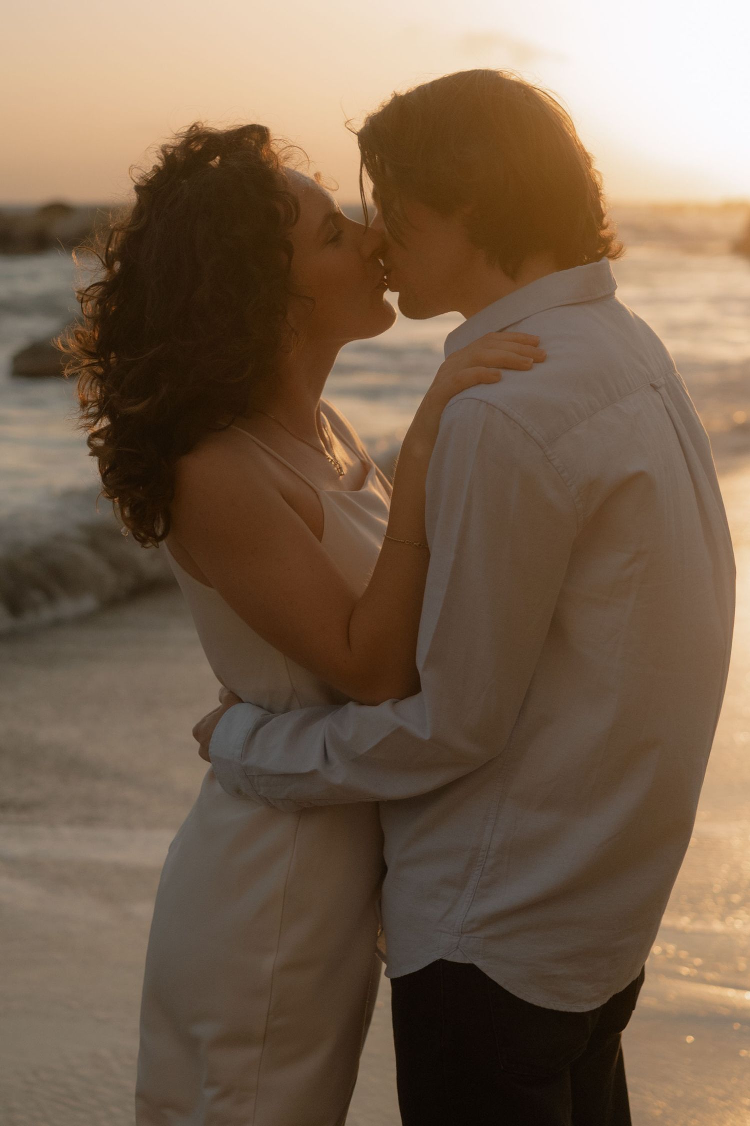 Couple sharing a romantic kiss on the beach at golden sunset, embracing each other by the ocean waves.